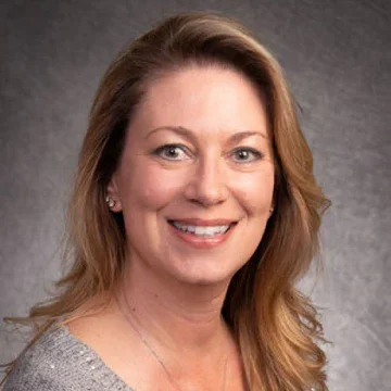 A woman with shoulder-length, light brown hair smiling at the camera against a gray background.