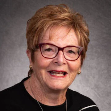 A smiling elderly woman with short reddish hair wearing glasses and a black top, against a gray background.