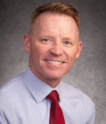 A man with short red hair, wearing a light blue dress shirt and a red tie, smiling against a gray textured background.