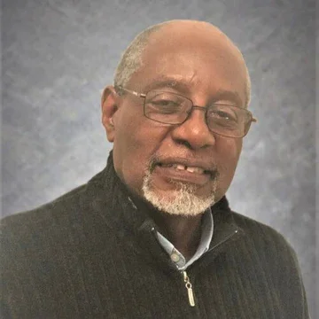 A smiling middle-aged man with glasses, gray hair, and a beard, wearing a dark zip-up sweater and standing against a gray background.