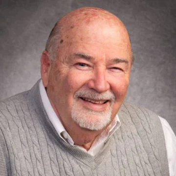 Portrait of an older man with a beard and bald head, smiling, wearing a gray sweater over a collared shirt.