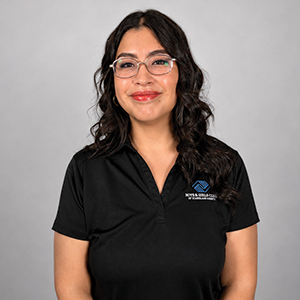 A woman with black hair, glasses, and a black collared shirt with a logo, smiling against a gray background.
