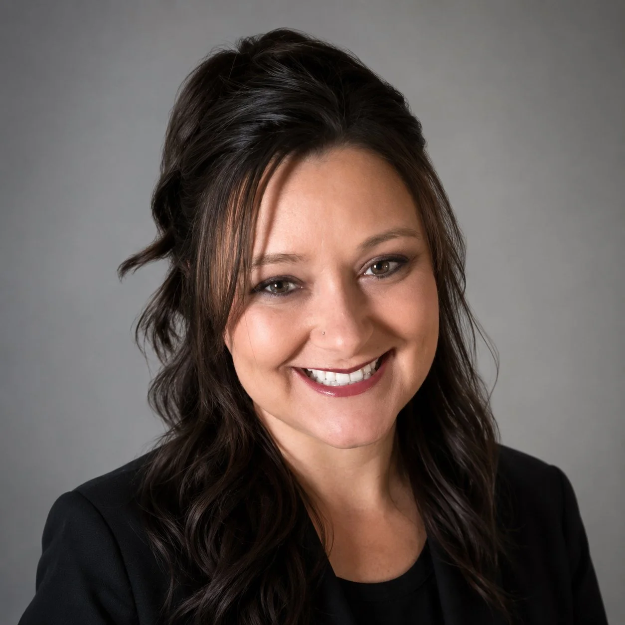 Portrait of a woman with long dark wavy hair, smiling, wearing a black blazer against a gray background.