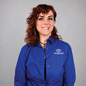 A woman with curly brown hair wearing a blue collared shirt with a logo, smiling at the camera against a light gray background.