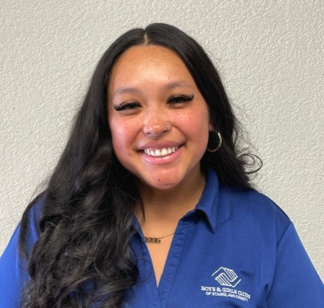 A young woman with long dark wavy hair, wearing a blue collared shirt with a logo that reads "Boys & Girls Club of Stambaugh-Courtney," smiling against a plain light-colored wall.