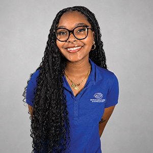 A young woman with long curly hair, glasses, and a bright smile, wearing a blue polo shirt with a logo, standing against a gray background.