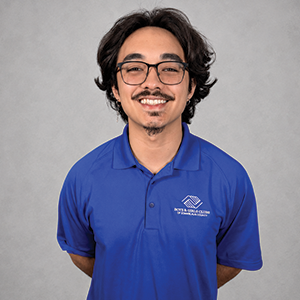 A young man with black wavy hair, glasses, and a mustache, smiling, wearing a blue polo shirt with a logo, standing against a plain gray background.