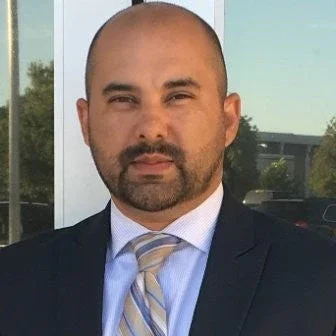 A man with a shaved head and beard wearing a dark suit, light blue shirt, and striped tie, standing outdoors in front of a window reflecting trees and sky.