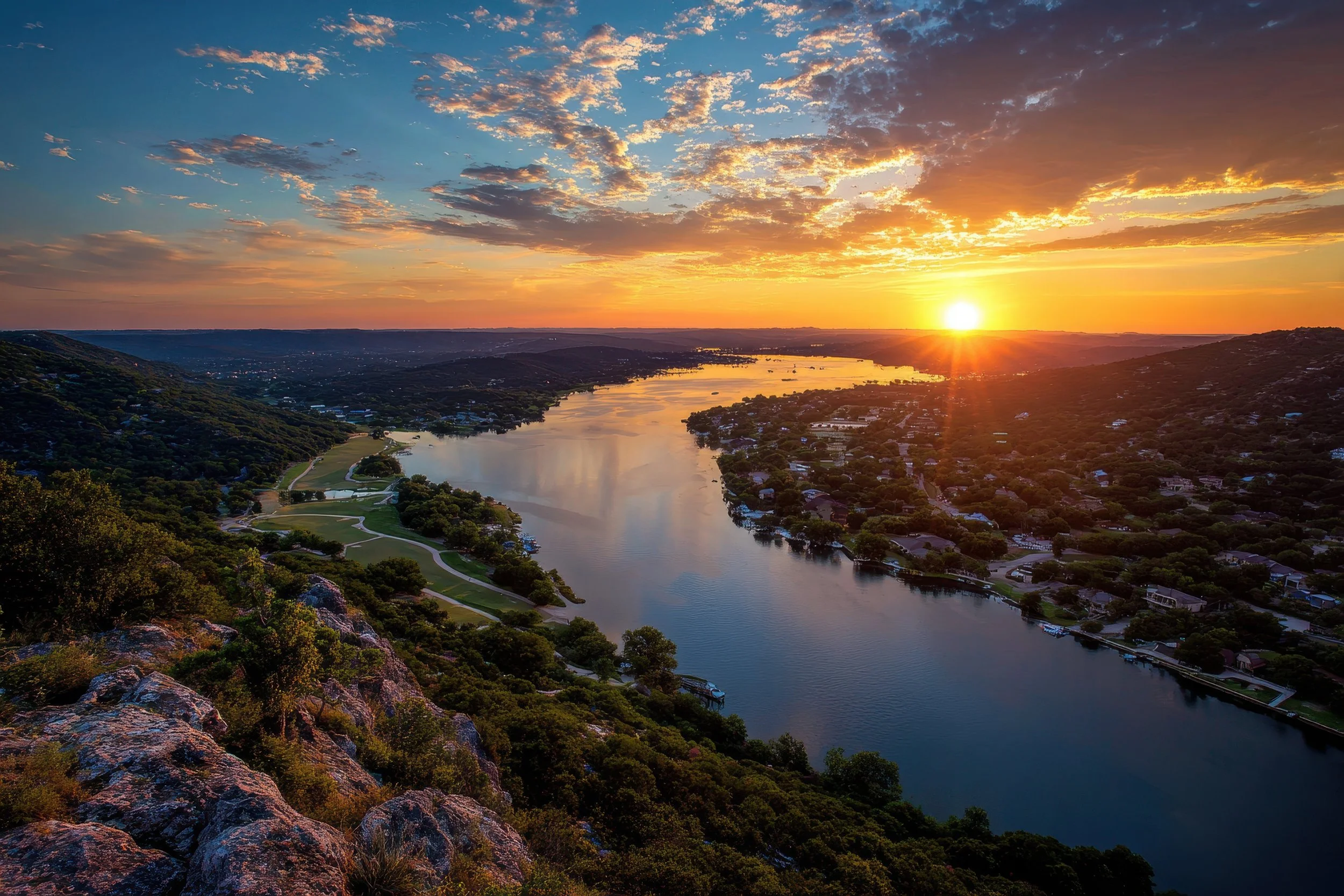 Sunset over a river with houses and green hills on both sides, with a partly cloudy sky.