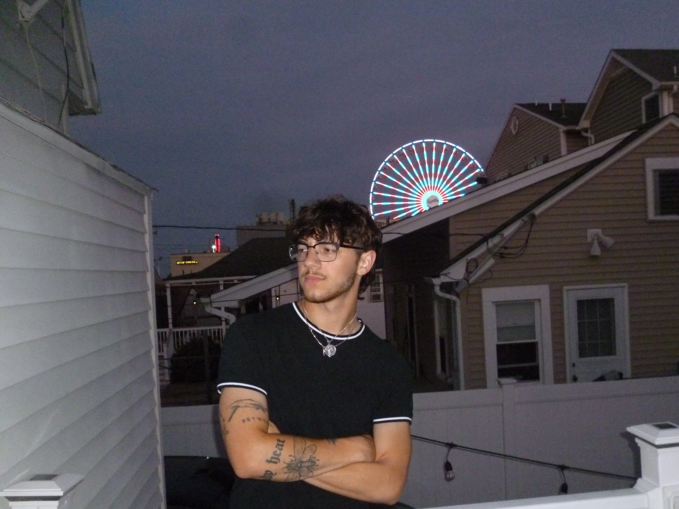 A young man with glasses, tattoos on his arms, wearing a black t-shirt, standing on an outdoor balcony or porch at night with a Ferris wheel illuminated in the background.