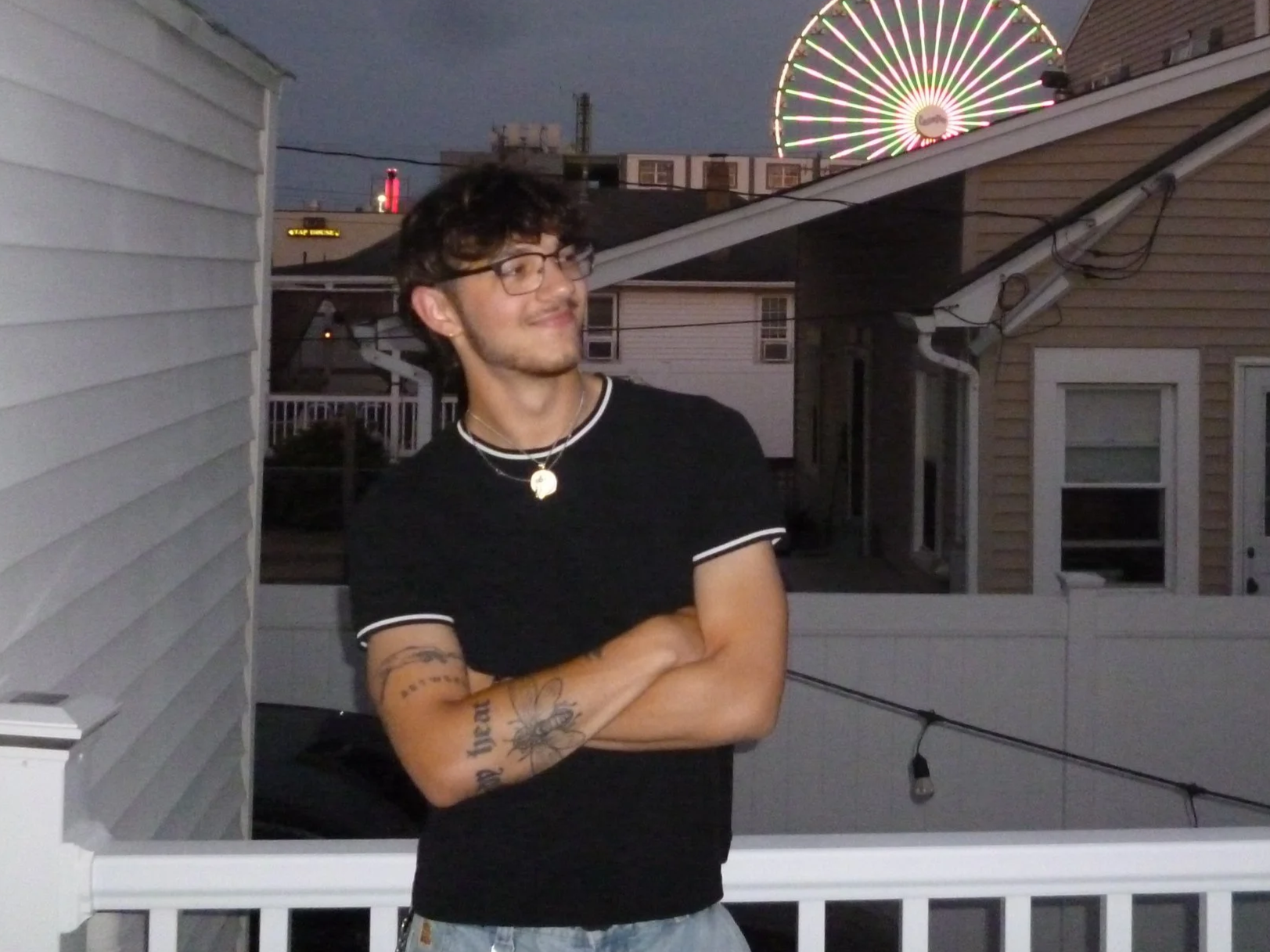 A young man with glasses and tattoos standing on a balcony at dusk, with a lit Ferris wheel in the background.