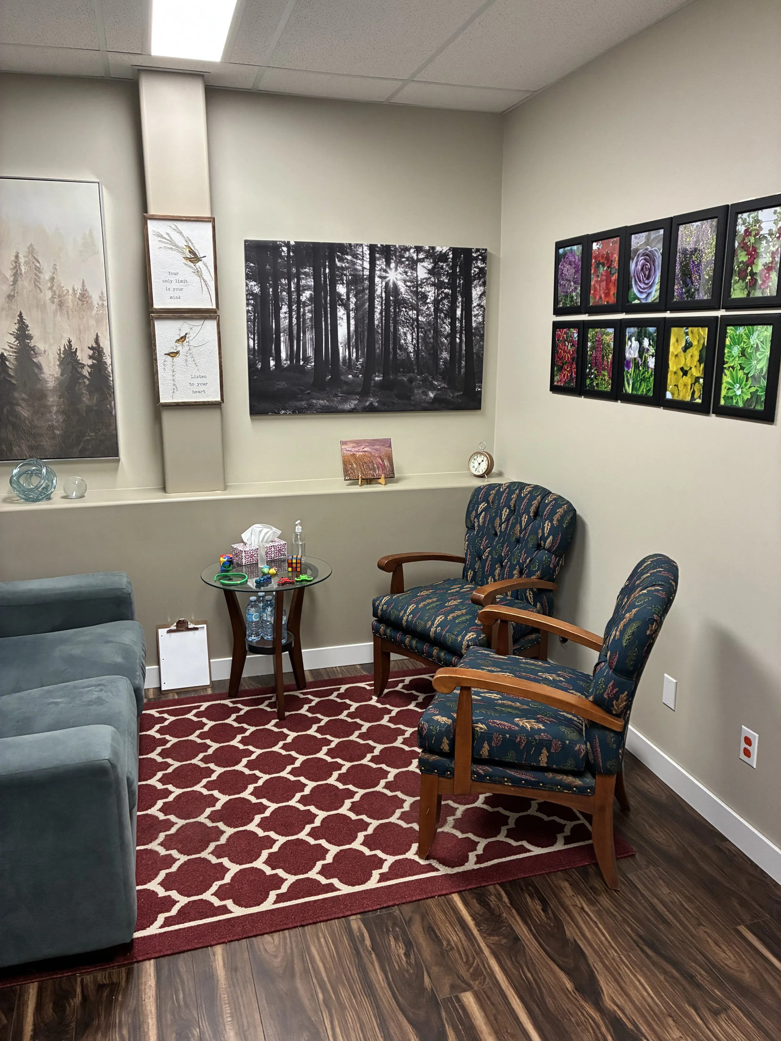 A cozy waiting room with two patterned armchairs and a cushioned sofa around a small glass table with toys and water bottles. The room has a red patterned rug, framed artwork of forest scenes and flowers on the walls, a clock, and a tissue box.