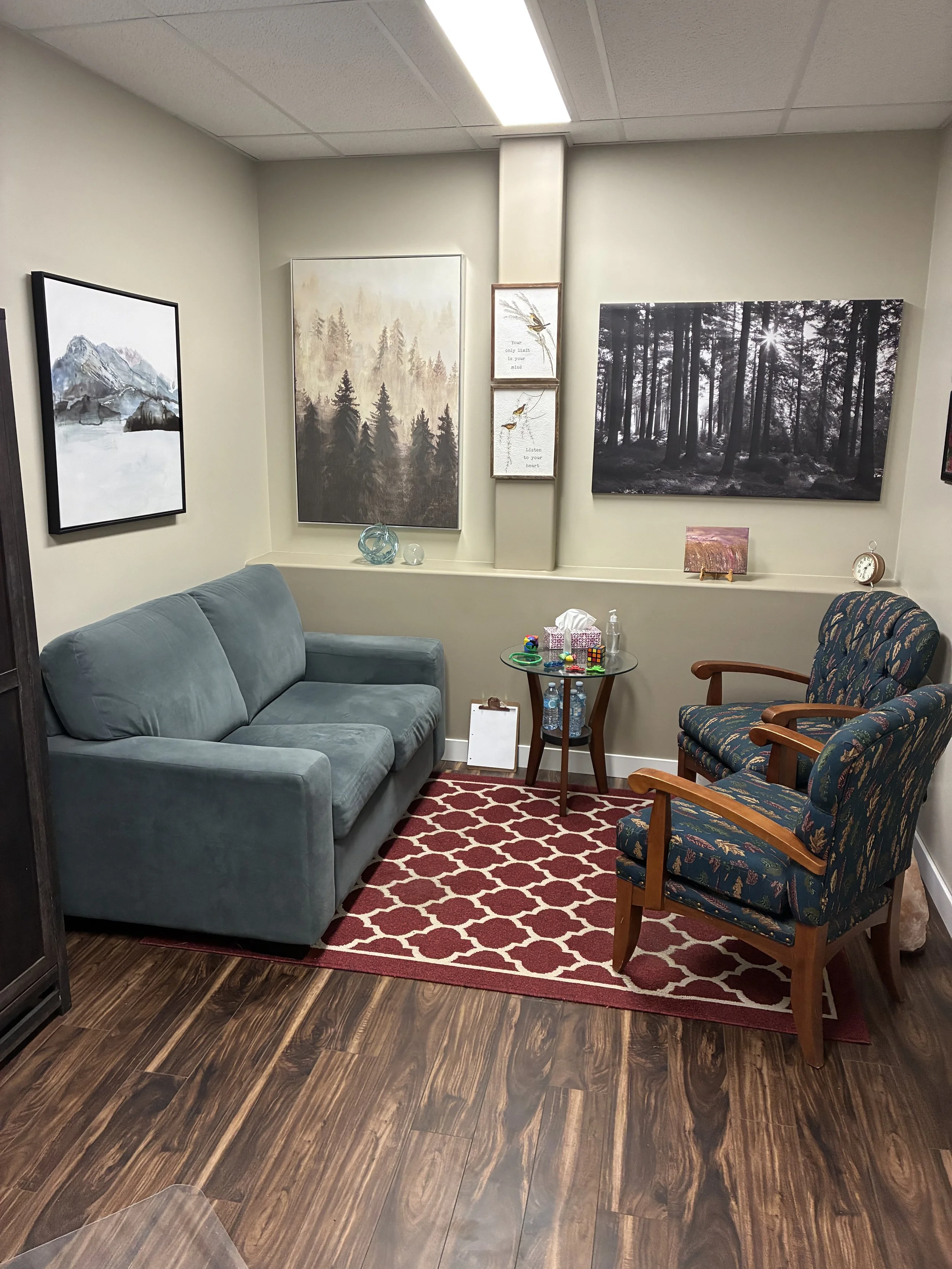 A cozy therapy office waiting room in Swift Current with a green sofa, patterned armchairs, and a small round table. The wall is decorated with framed photographs and artwork of mountains and forests. The floor is wood with a red patterned rug.