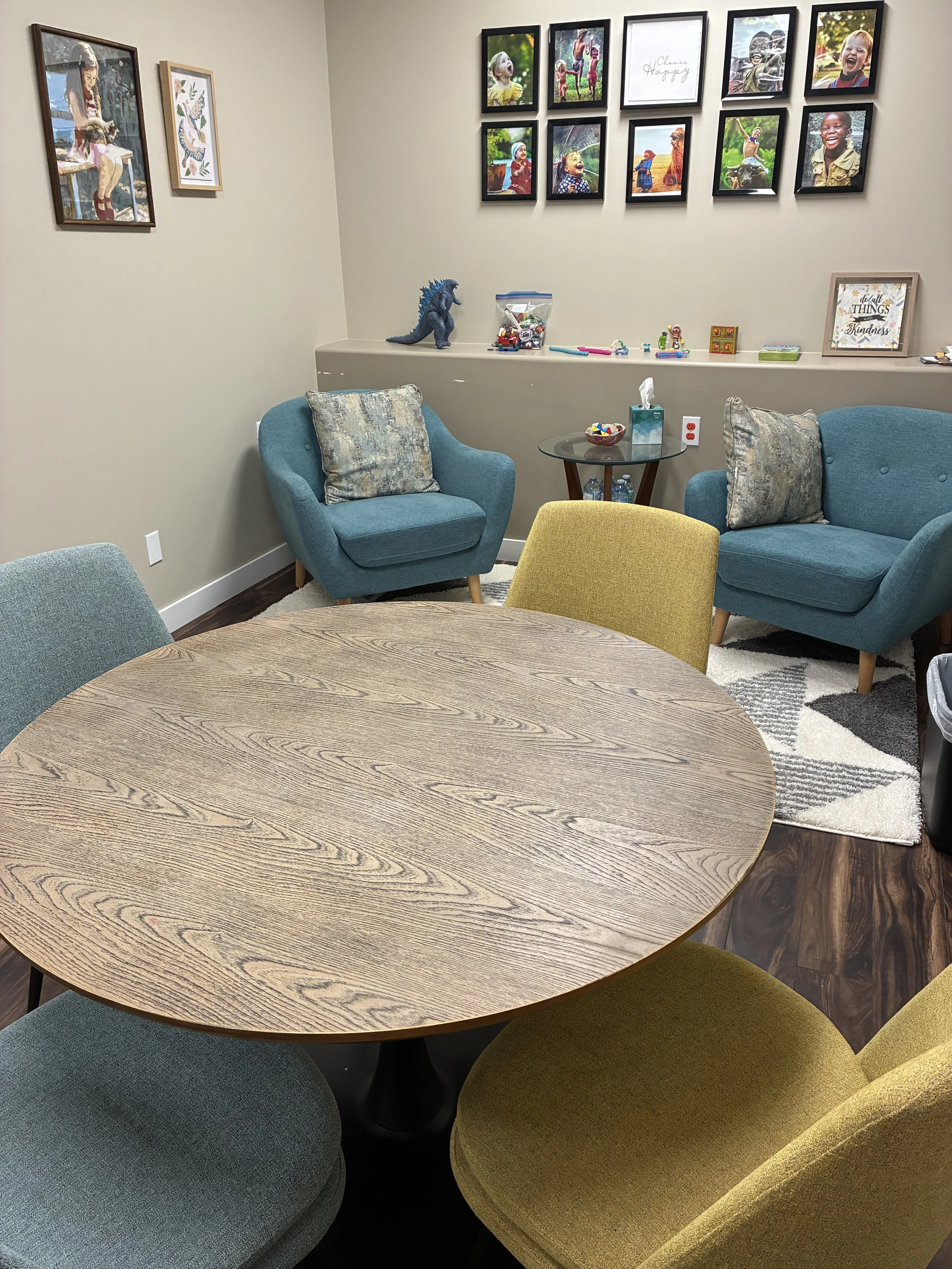 A cozy waiting room with a round wooden table surrounded by colorful upholstered chairs, a blue armchair, a teal armchair, and a yellow chair. In the background, a wall displays a gallery of framed photographs of children laughing and playing, along with decorative artwork and framed quotes. There is a side table holding toys and water bottles, and a patterned rug on the dark wooden floor.