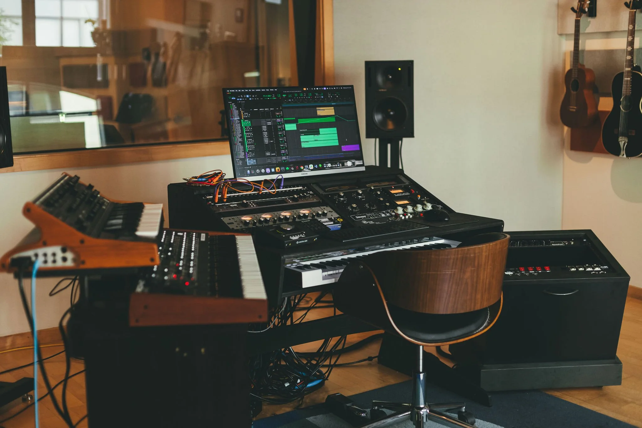 Music production studio with two guitars hanging on the wall, a computer monitor displaying digital audio workstation software, a mixing console, keyboard, and various electronic musical instruments and equipment.