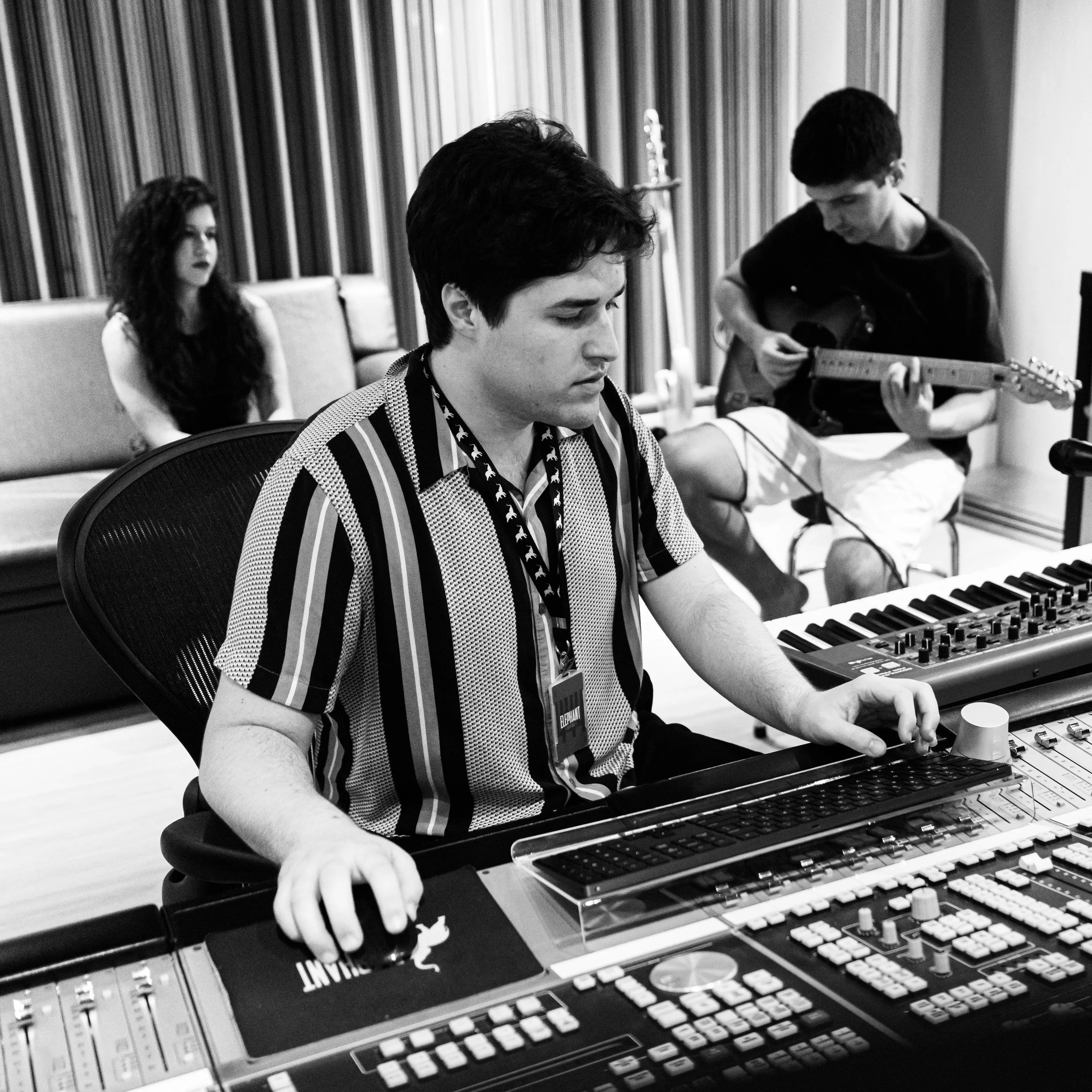 Black and white photo of a professional, high-level recording studio. One person is operating an SSL console, another is holding a guitar, and the third is watching the audio-professional