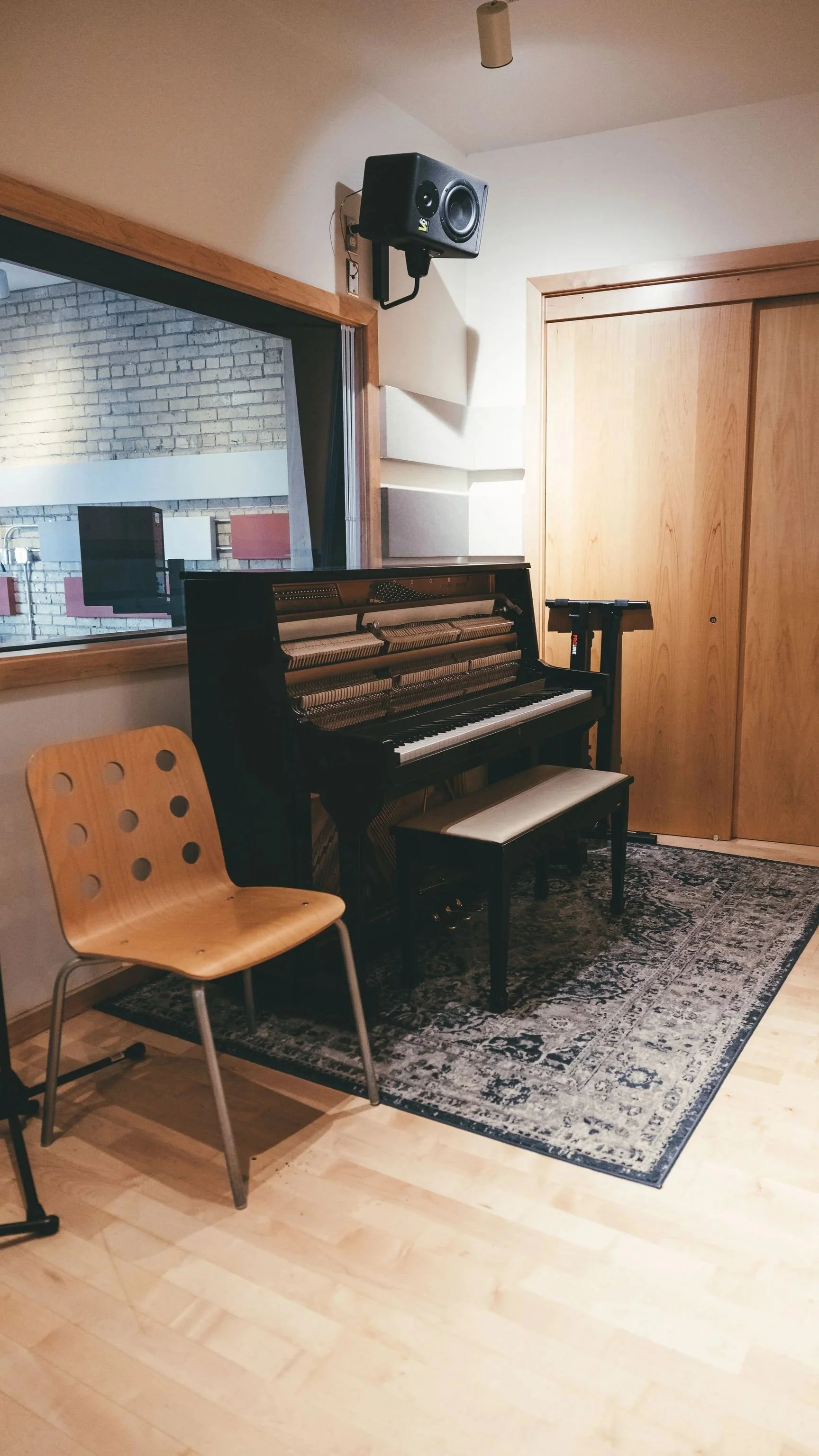 An indoor room with a black upright piano on a patterned rug, a wooden chair, a music stand, and a wooden door. There is a large window showing an exterior brick wall and part of a studio.