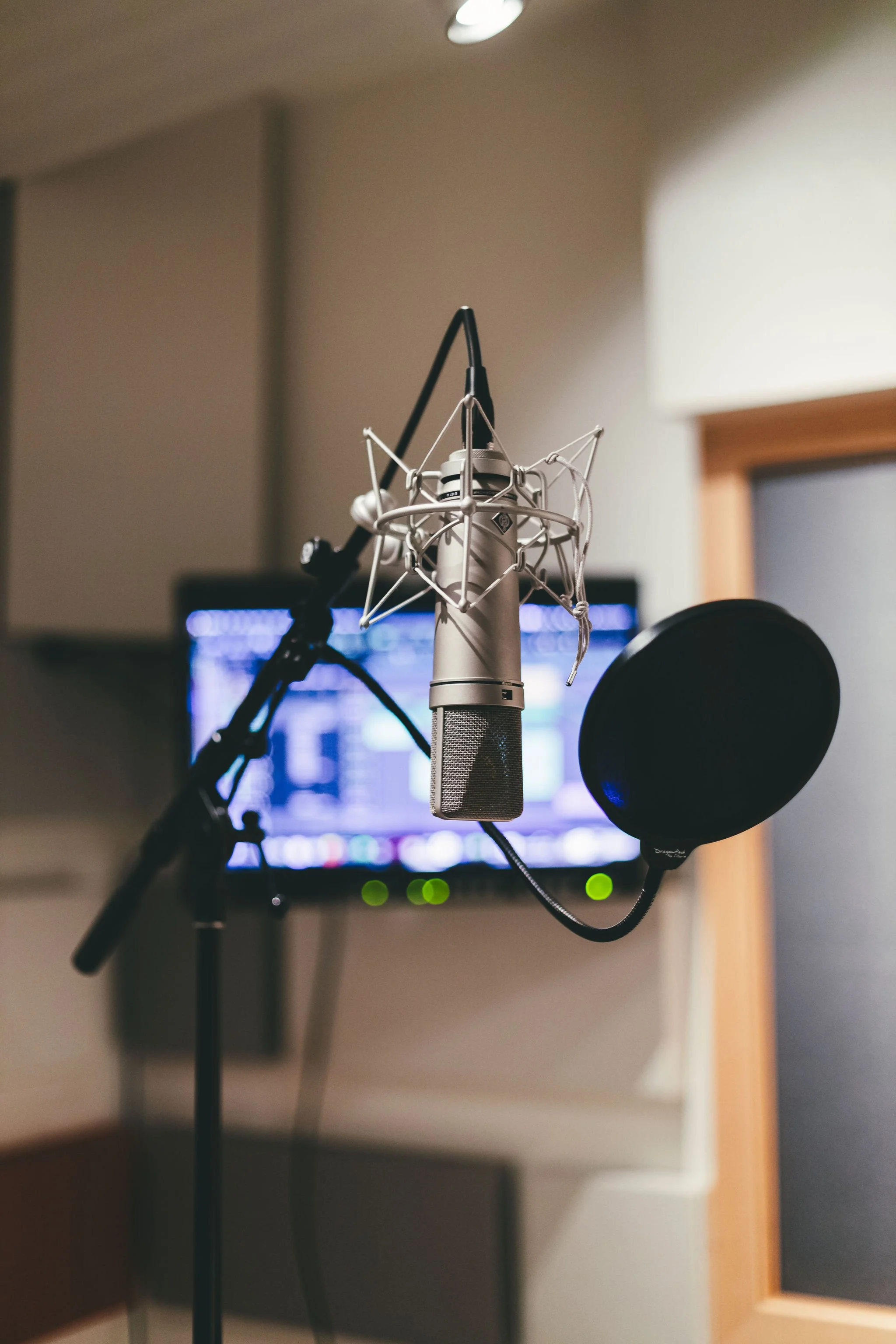 A professional microphone with pop filter in a recording studio, with computer screens and sound equipment in the background.