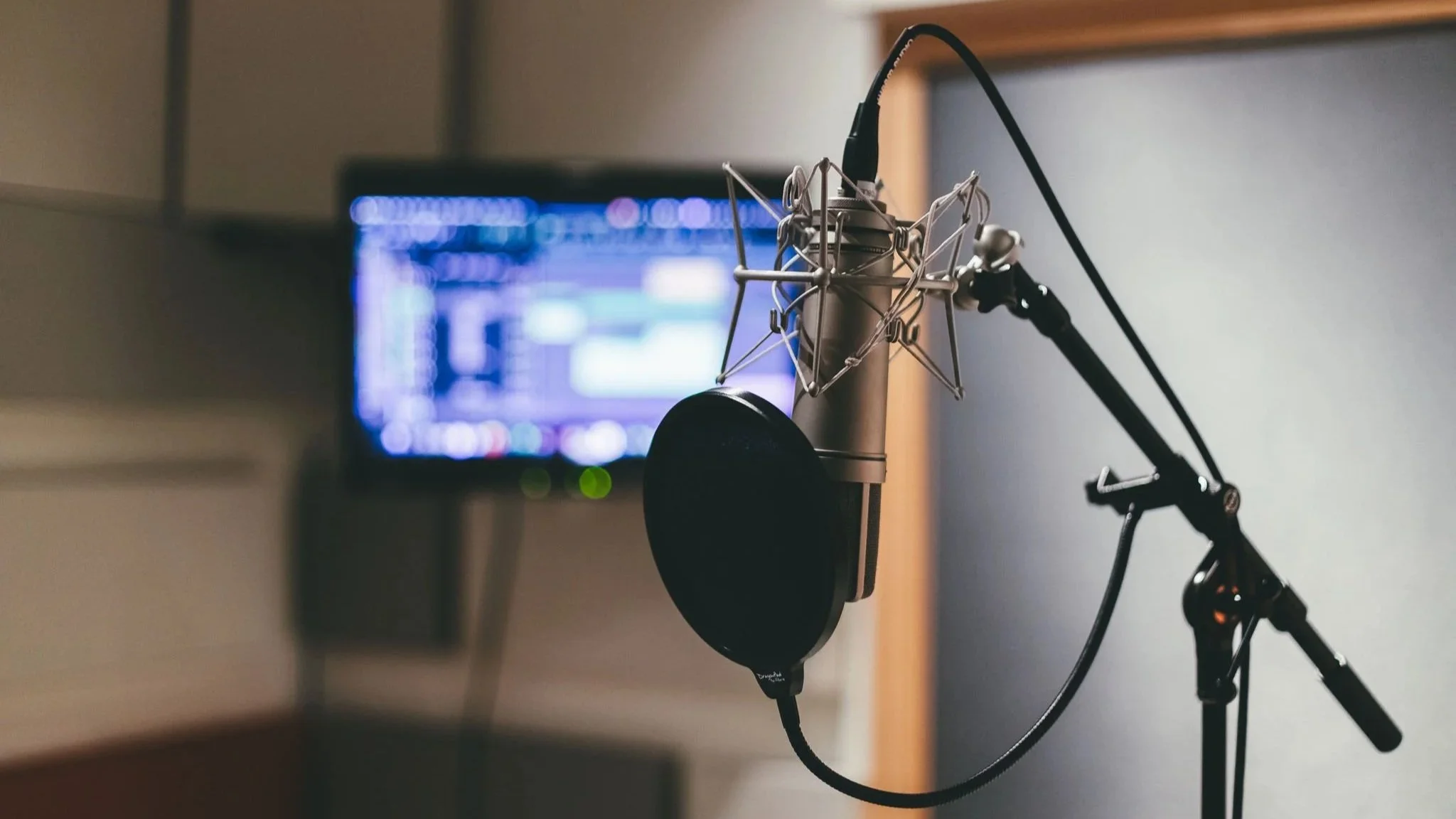 A microphone with a pop filter is set up in a recording studio, with a computer monitor displaying audio software in the background.