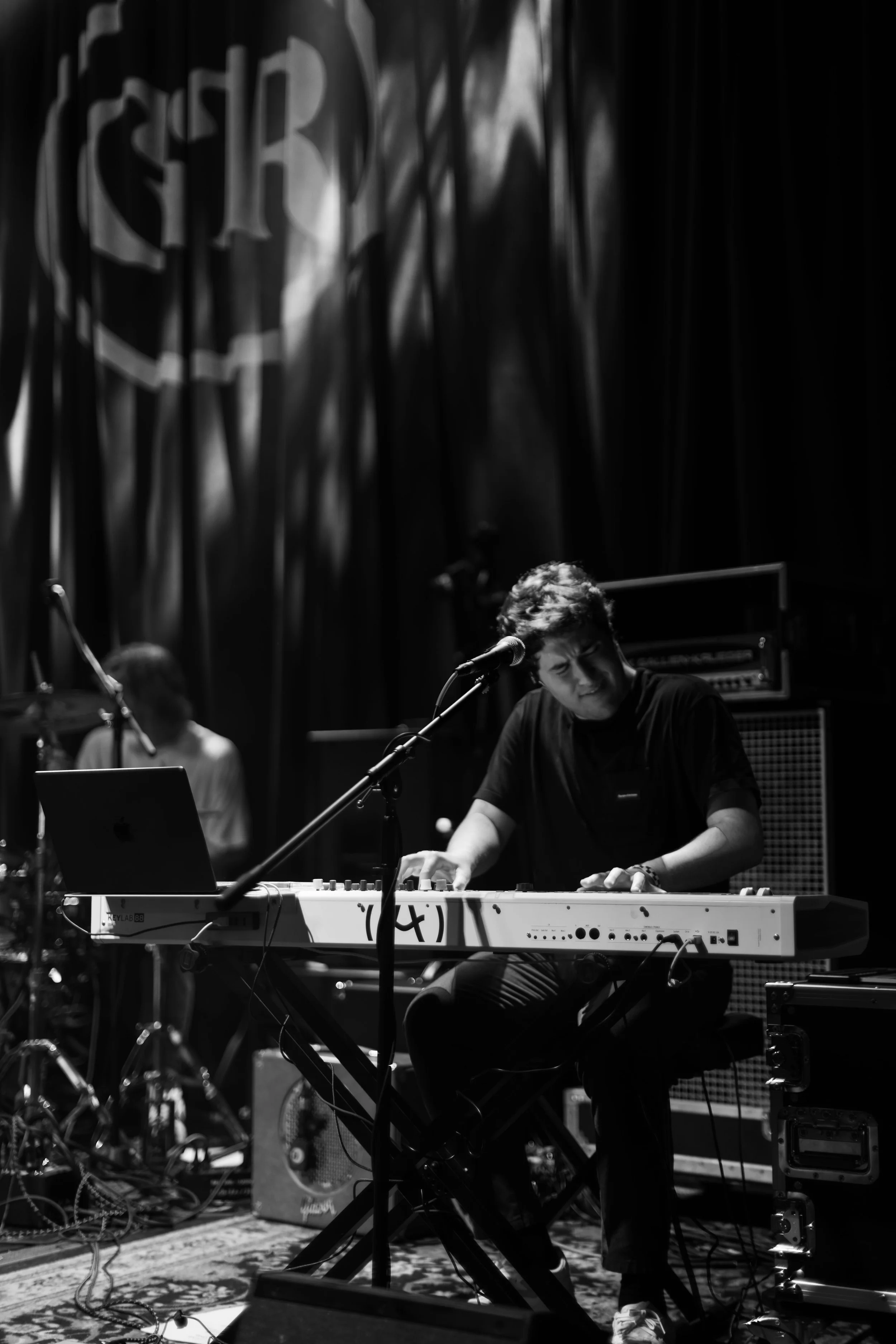 A musician playing a keyboard on stage during a live performance, with a person in the background and stage equipment visible.