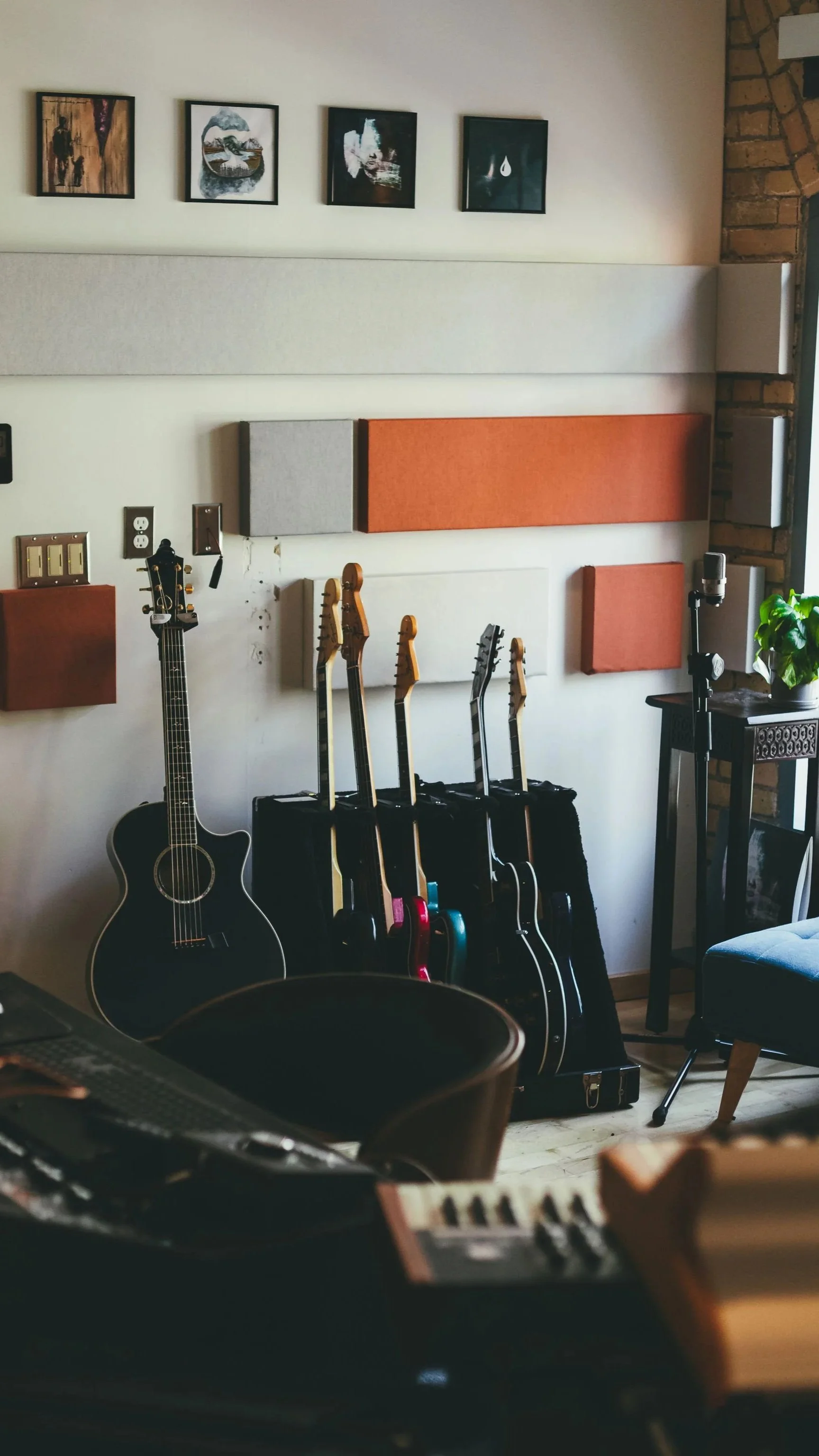A music studio with guitars hanging on a wall, acoustic panels, and a microphone, with a plant on a side table and framed artwork on the wall.