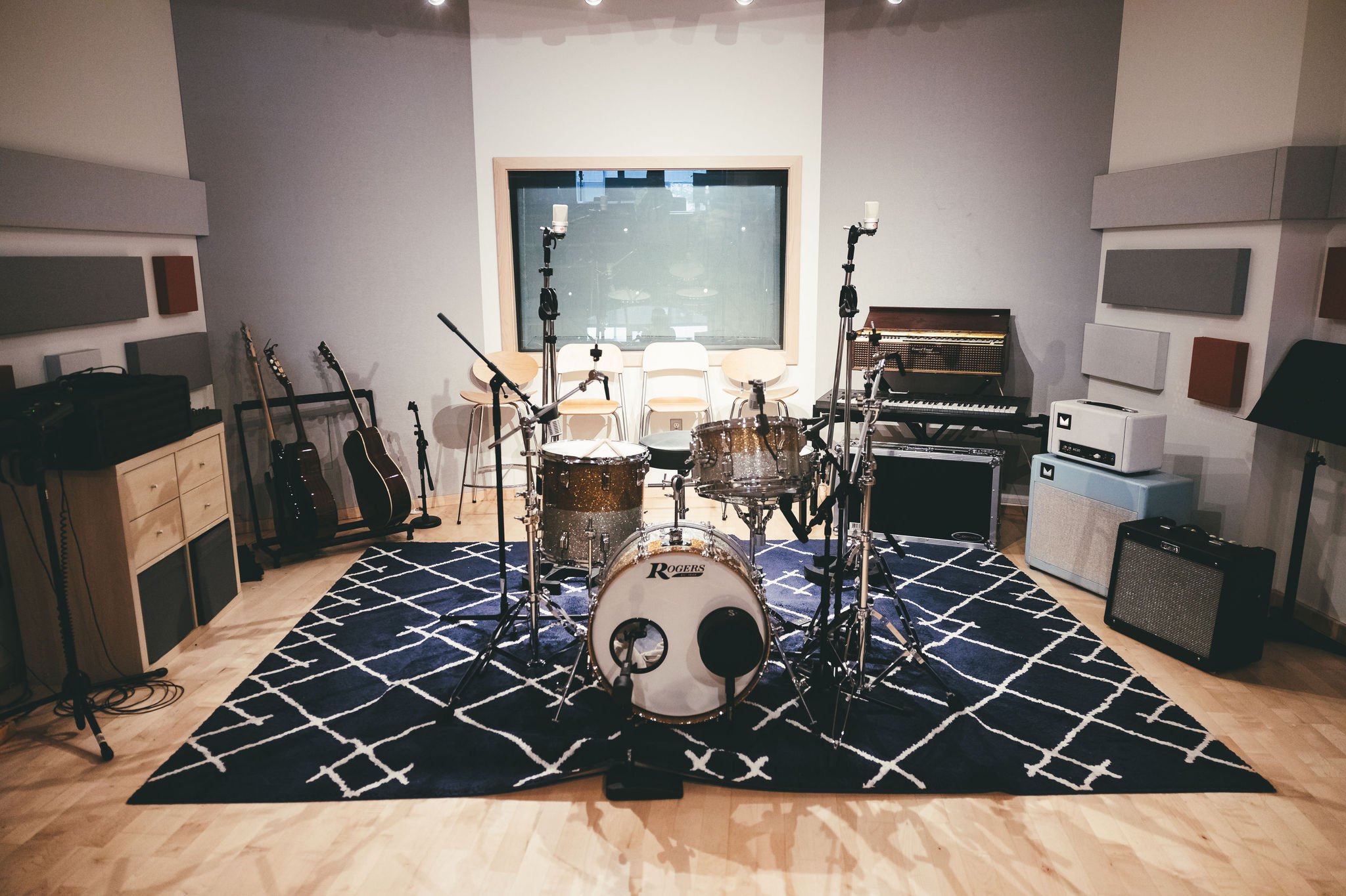 Music studio with drums, guitars, microphones, a keyboard, amps, and acoustic panels, all set up for recording or practicing. The room has a black and white geometric rug and a large window at the back.