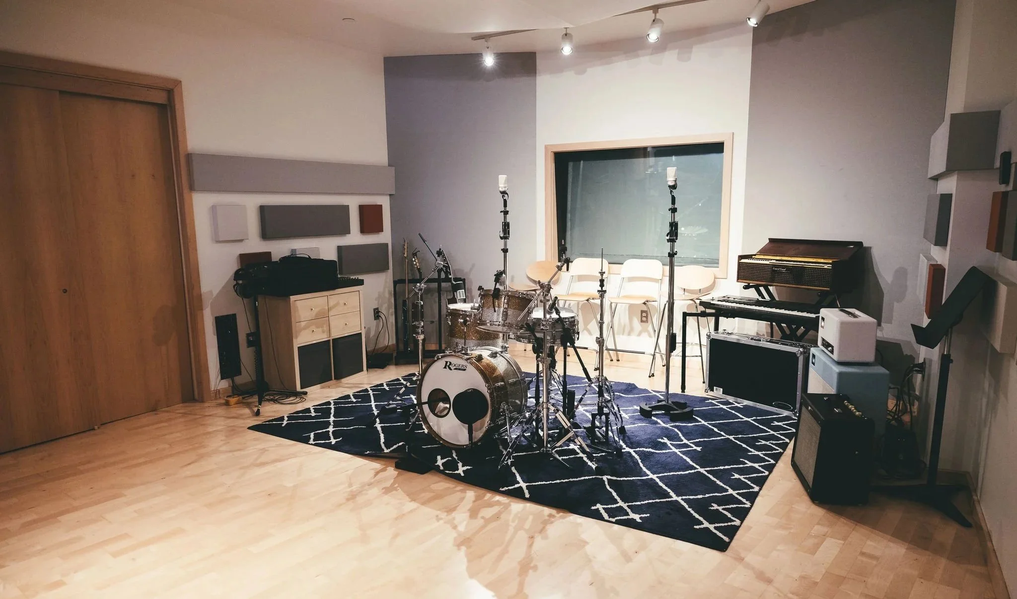 Music studio with drum set, keyboard, guitar, and seating area, decorated with acoustic panels and in-ceiling track lighting.