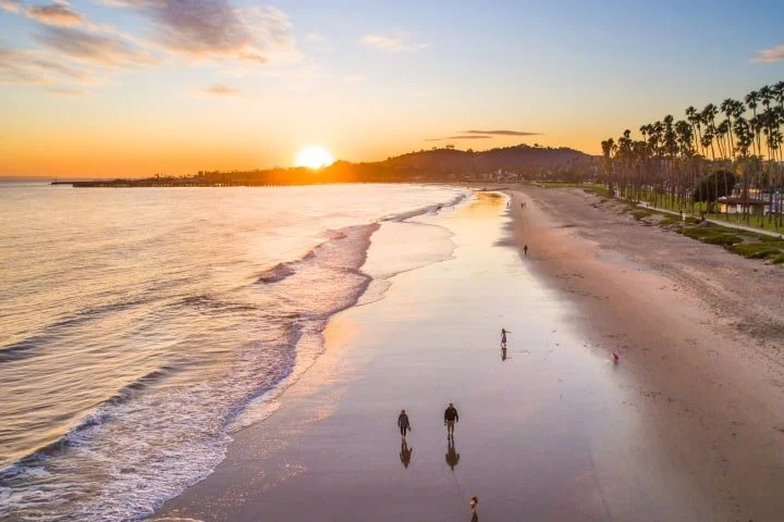 People walking on the wet sand of a beach during sunset, with the ocean on the left and palm trees along the shoreline on the right.