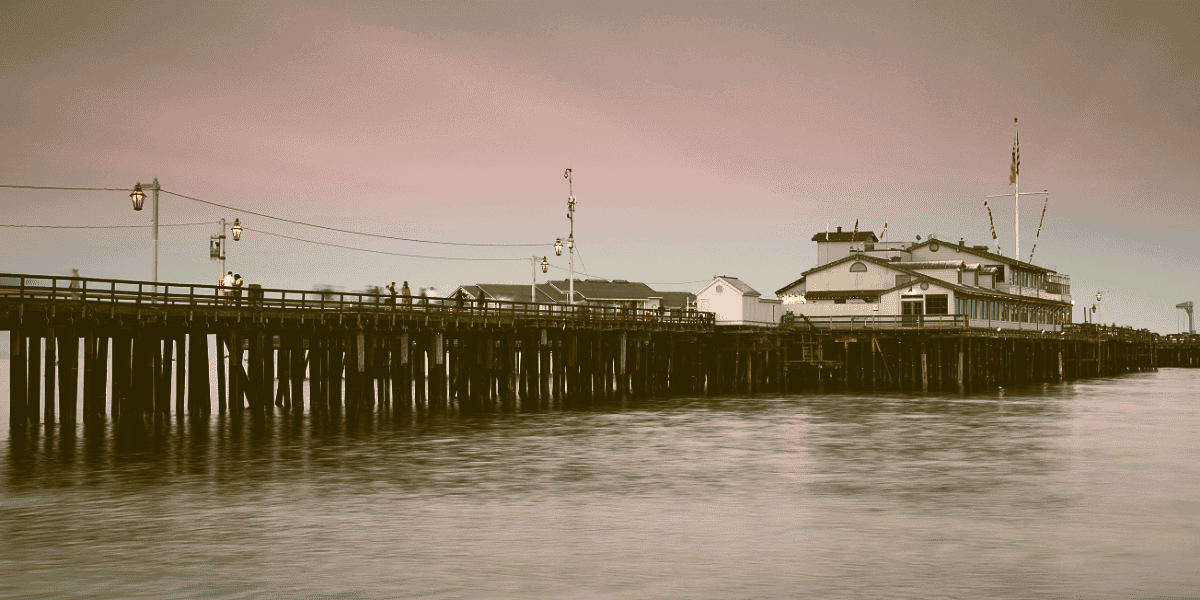 A wooden pier extending over calm water with a large building at the end, small houses along the pier, and a flag on the building, all under a dusky sky.