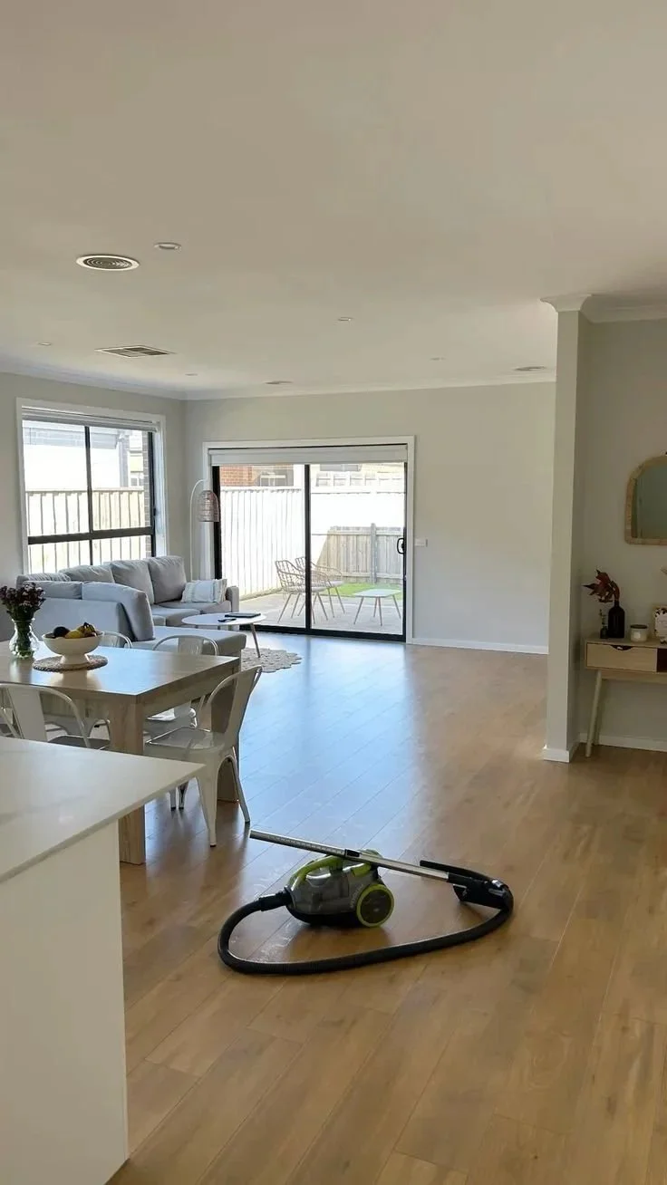 Salon lumineux avec canapé gris, table basse, ouverture sur patio avec deux chaises, aspirateur sur le sol en bois.
