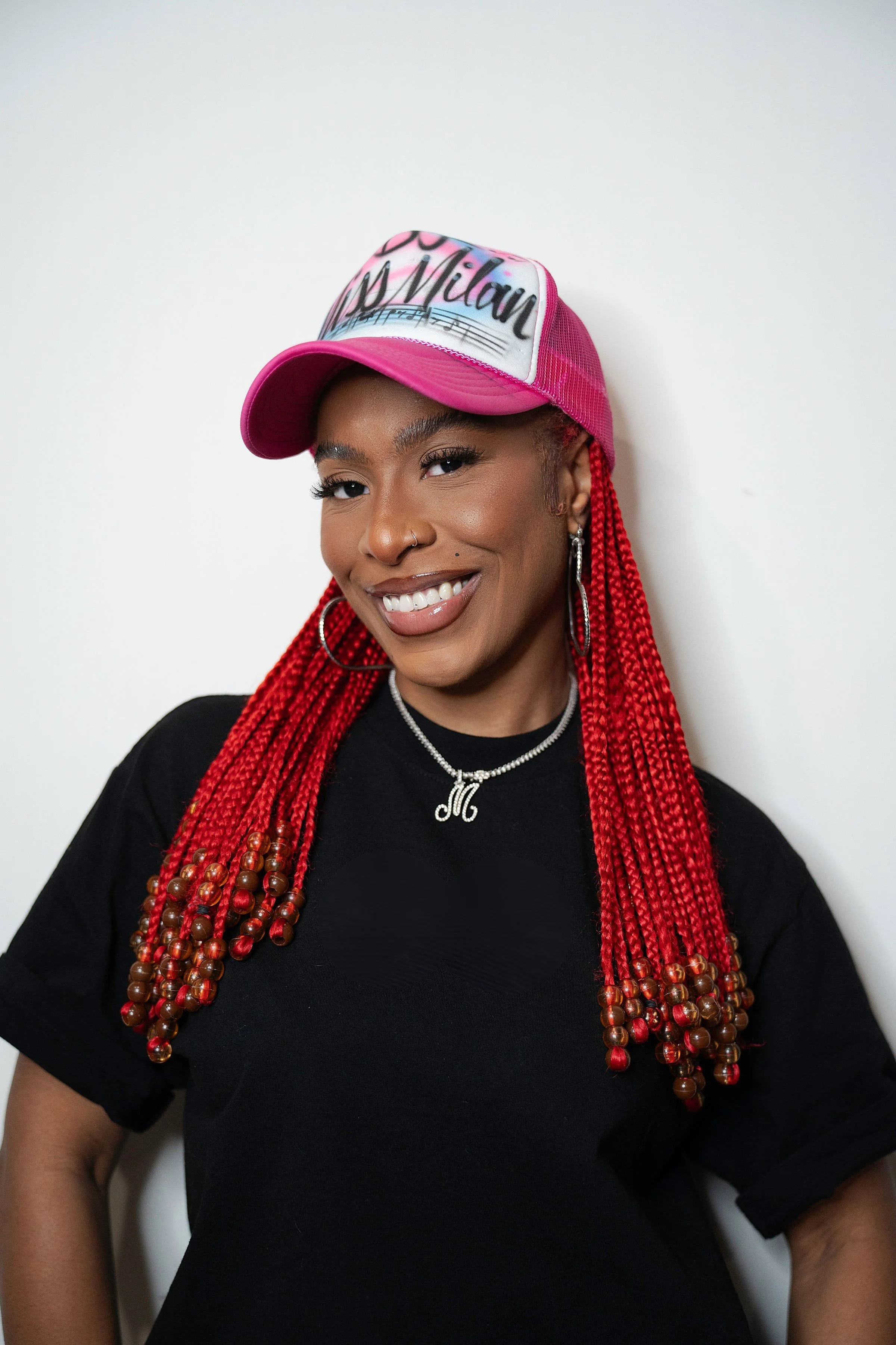 A woman with long red braided hair wearing a pink and white baseball cap with the name 'Milan' on it, hoop earrings, a necklace, and a black t-shirt, smiling at the camera against a plain white background.