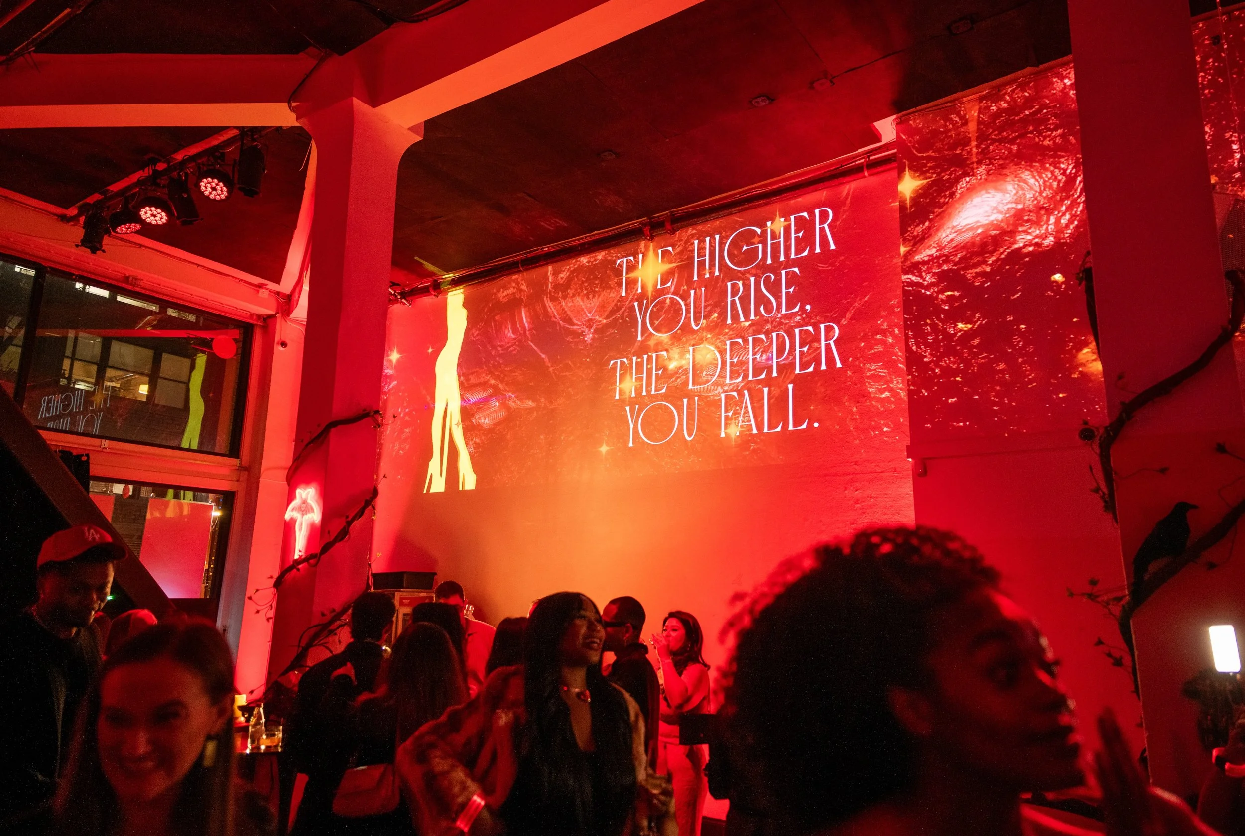 A crowded indoor event with red lighting, featuring a large digital screen displaying the message "The higher you rise, the deeper you fall." There are people of diverse ages and genders, some taking photos, and decorative string lights hanging from 