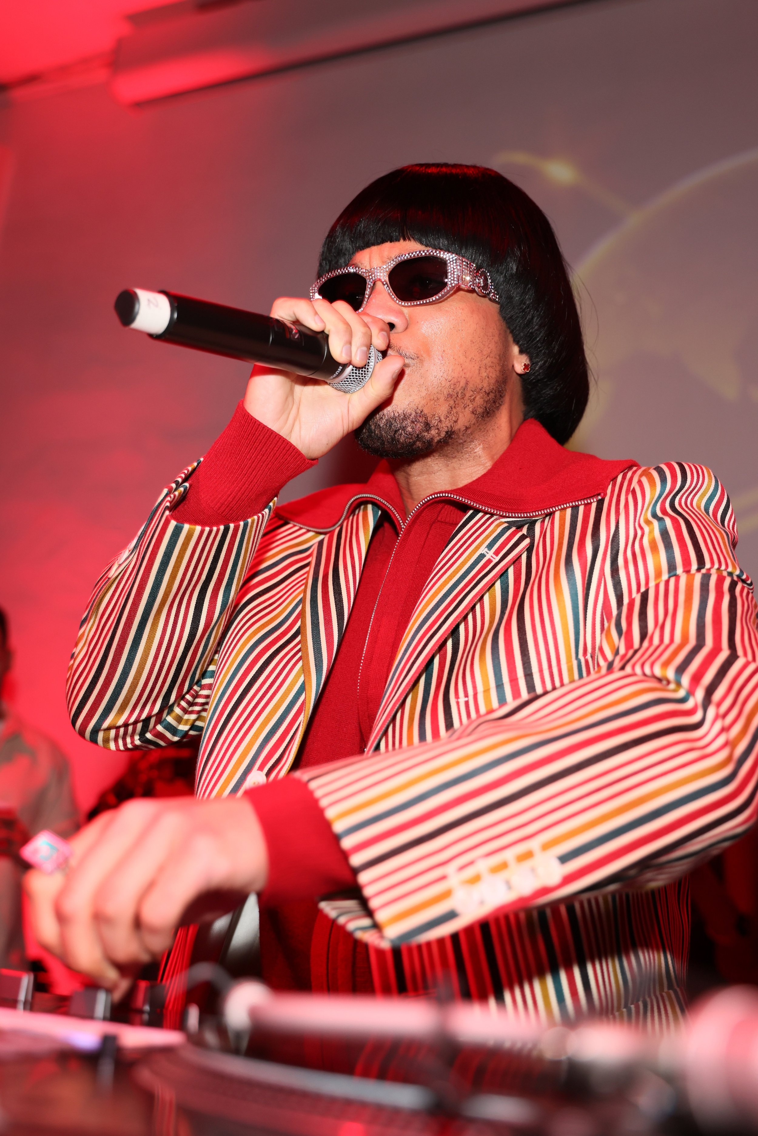 Person DJ wearing black wig, rhinestone sunglasses, red jacket, and striped blazer playing music with DJ equipment in a red-lit environment.