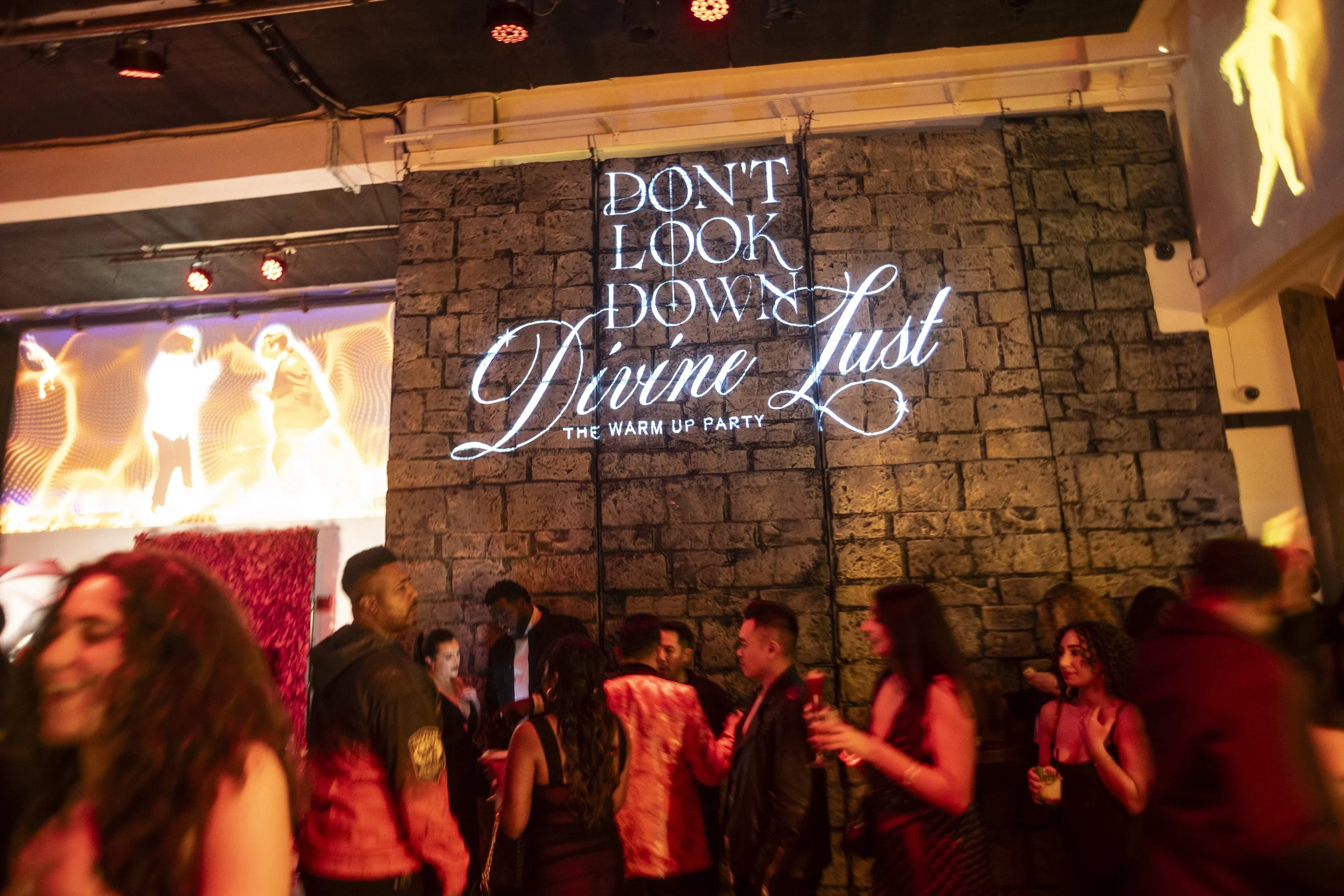 Crowd of people at a lively party or nightclub with a neon sign on a brick wall that reads 'Don't Look Down, Divine Just - The Warm Up Party.' People are socializing and enjoying the event.