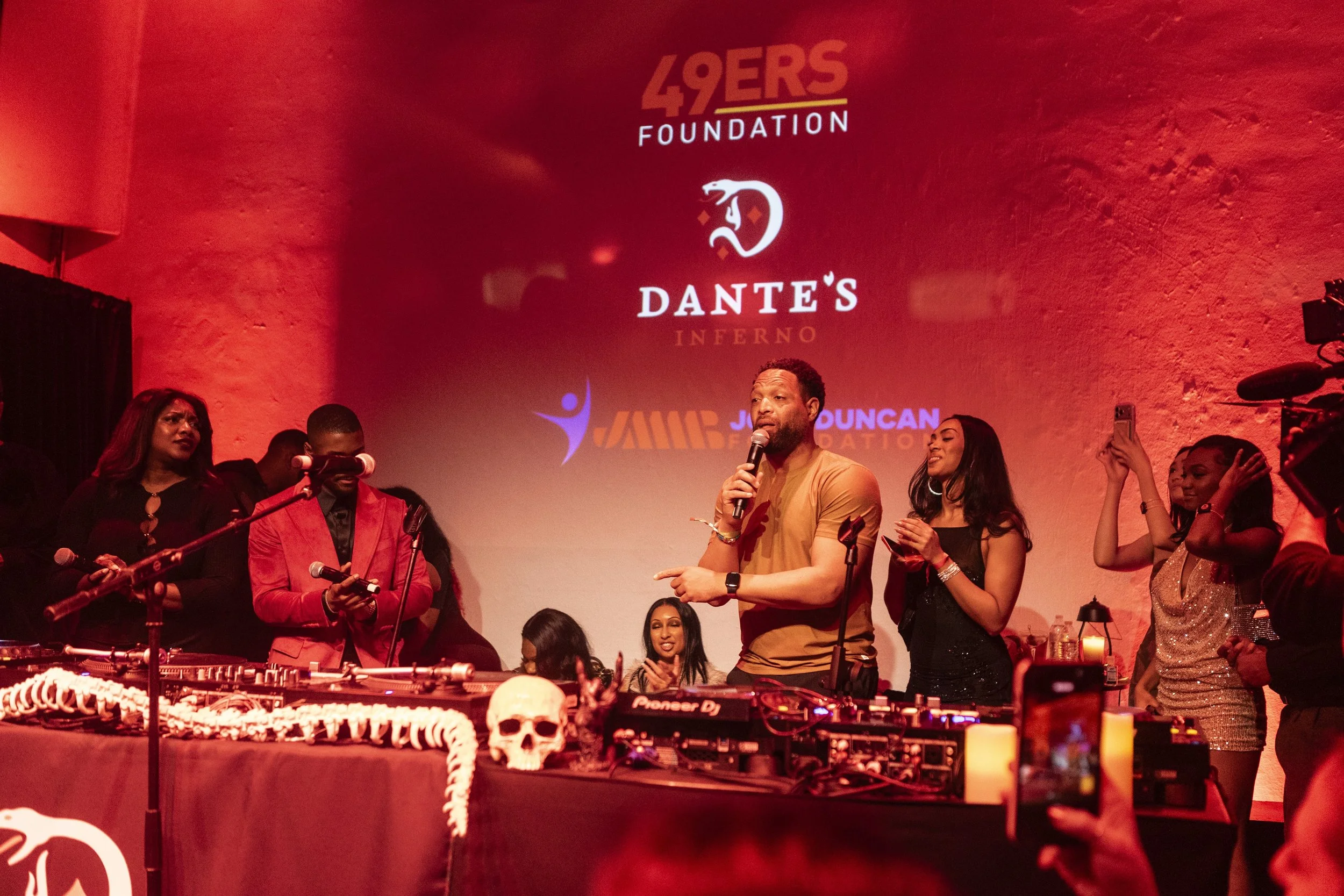 A group of diverse people at a party or event with a DJ setup and a skull decoration, standing in front of a red wall with logos and text, including '49ERS FOUNDATION' and 'DANTE'S INFERNO'.