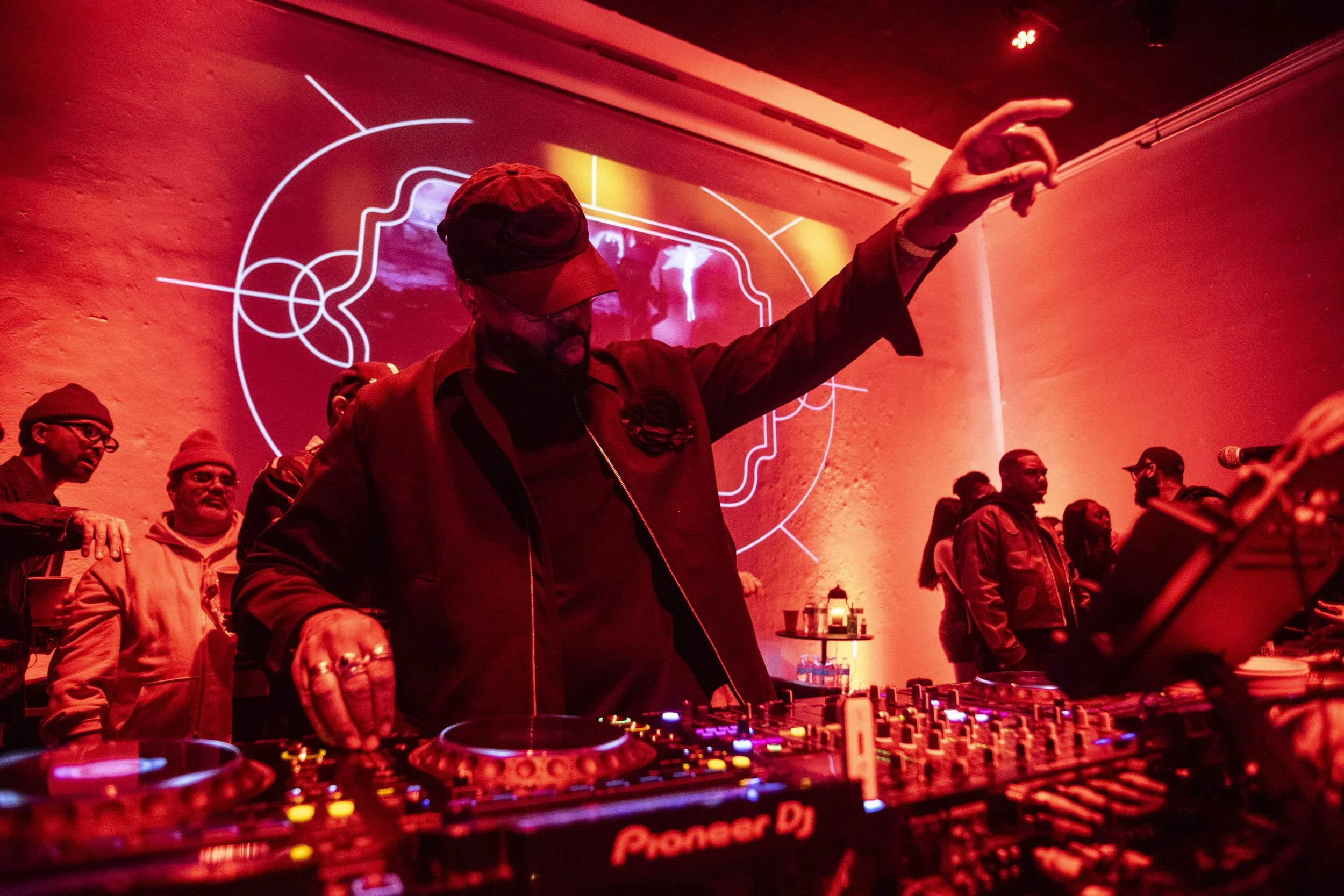 A DJ performing at a party or club, surrounded by people. The DJ is wearing a jacket and a cap, with one hand extended. The scene is lit with red lighting, and a neon-style graphic is visible on the wall behind him. DJ equipment, including a Pioneer 