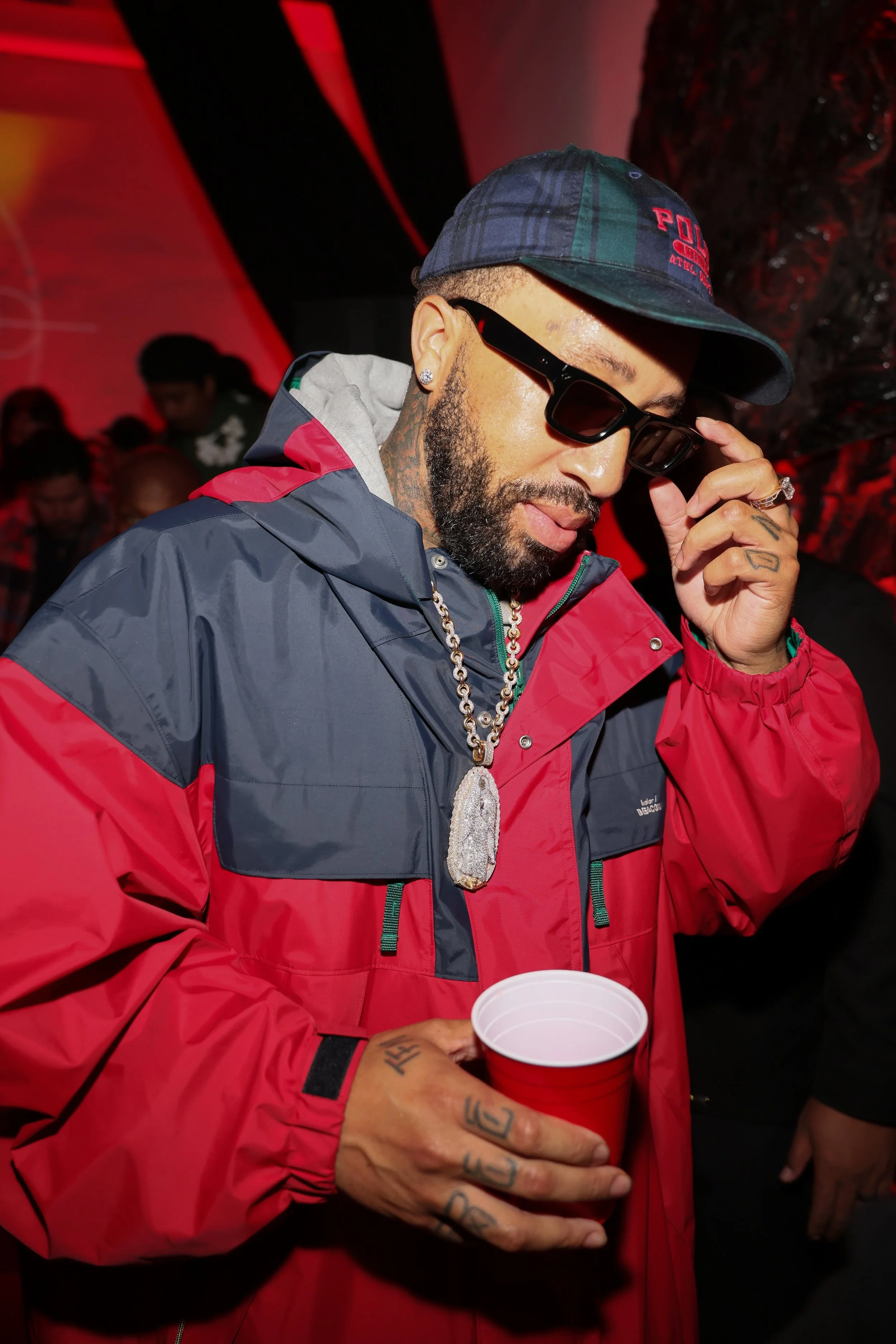 Close-up of a man wearing a blue plaid cap with red embroidery, sunglasses, and a red and gray jacket. He has tattoos on his hands and is holding a red plastic cup. The background is dimly lit with red lighting, and other people can be seen in the ba