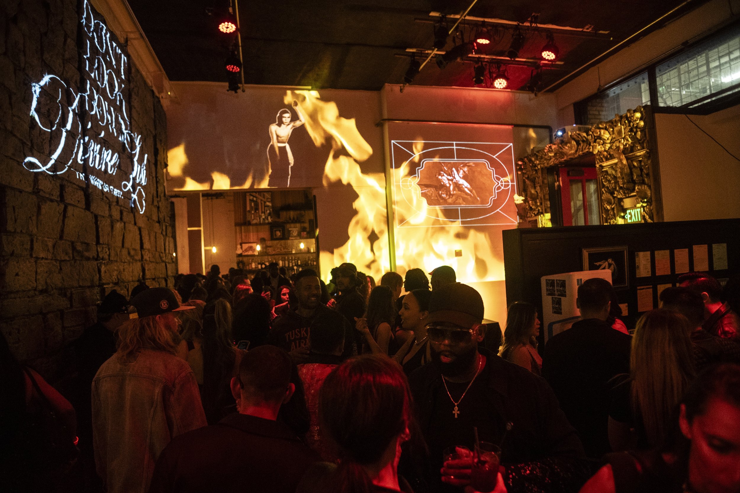 A crowded indoor event with people watching a stage with flames and projected images of a woman in black and white. The background includes brick walls, decorative elements, and a neon sign.