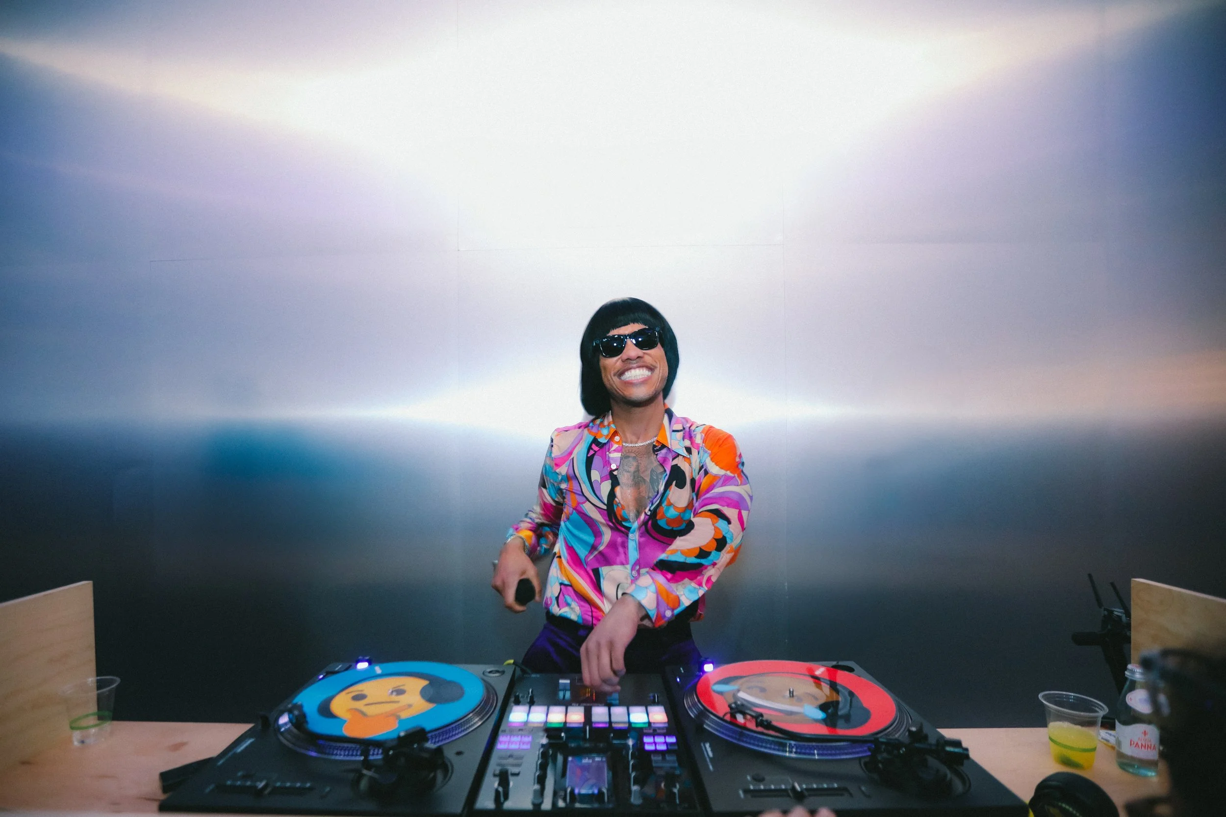 Person with dark hair and sunglasses smiling while DJing at a party, wearing a colorful patterned shirt surrounded by DJ equipment on a wooden table, with a plain wall in the background.
