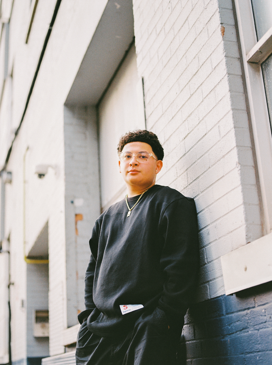 A young man with curly hair and glasses wearing a black sweatshirt and gold chain necklace, leaning against a white brick wall on an urban street.