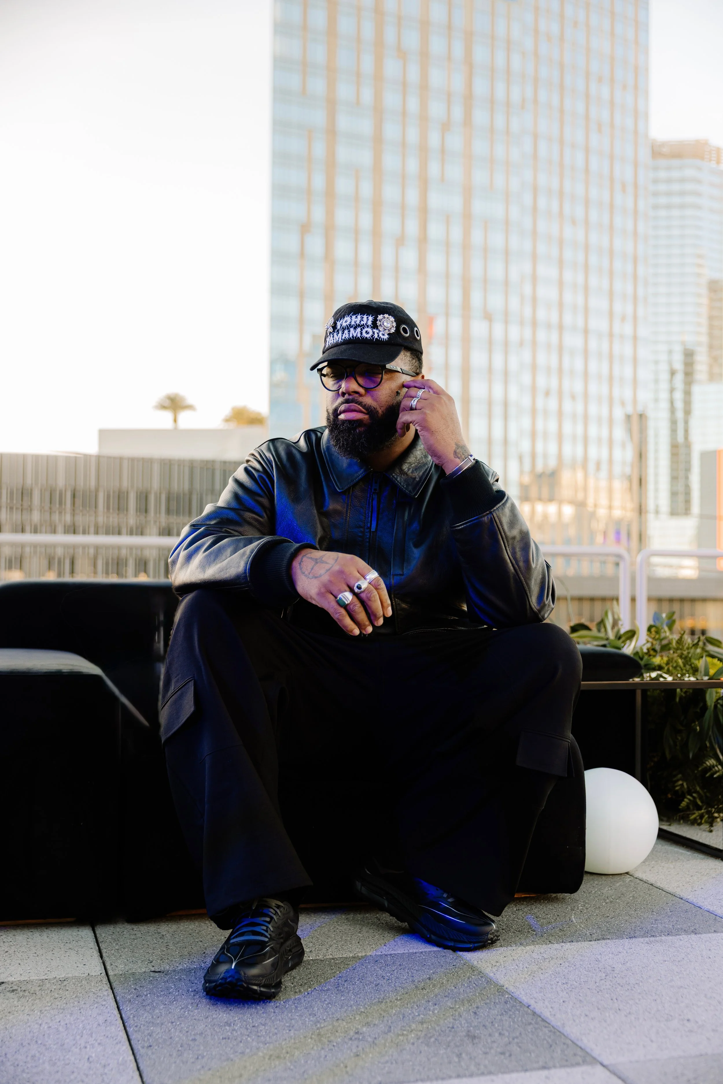 A man with a beard and glasses wearing a black leather jacket, black cap with decorative pins, sitting on a bench in an urban setting with large glass buildings in the background. He is touching his face with his right hand, looking thoughtful.