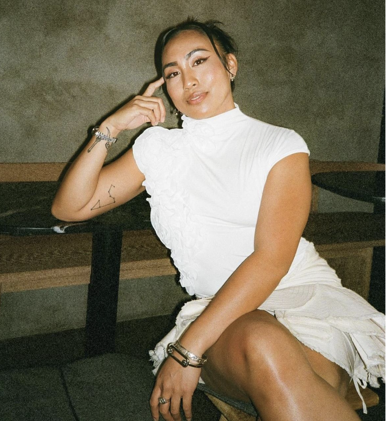 A woman with dark hair and light makeup sitting at a table in a white ruffled top and a cream skirt with a tattoo on her arm and various jewelry, posing with her hand near her head in an indoor setting.