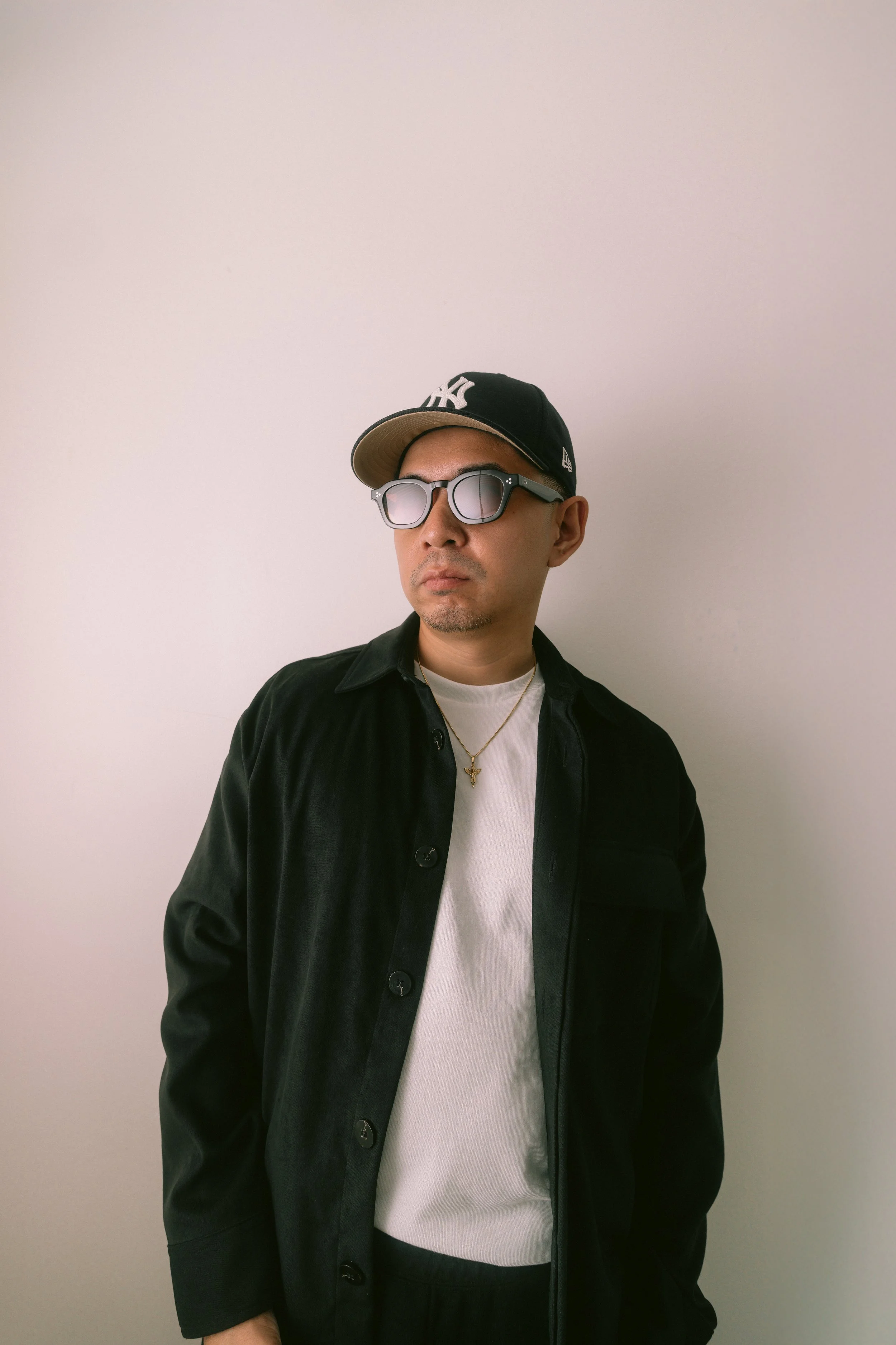 Man wearing a NY Yankees baseball cap, sunglasses, and a black jacket over a white shirt, posing against a plain wall.