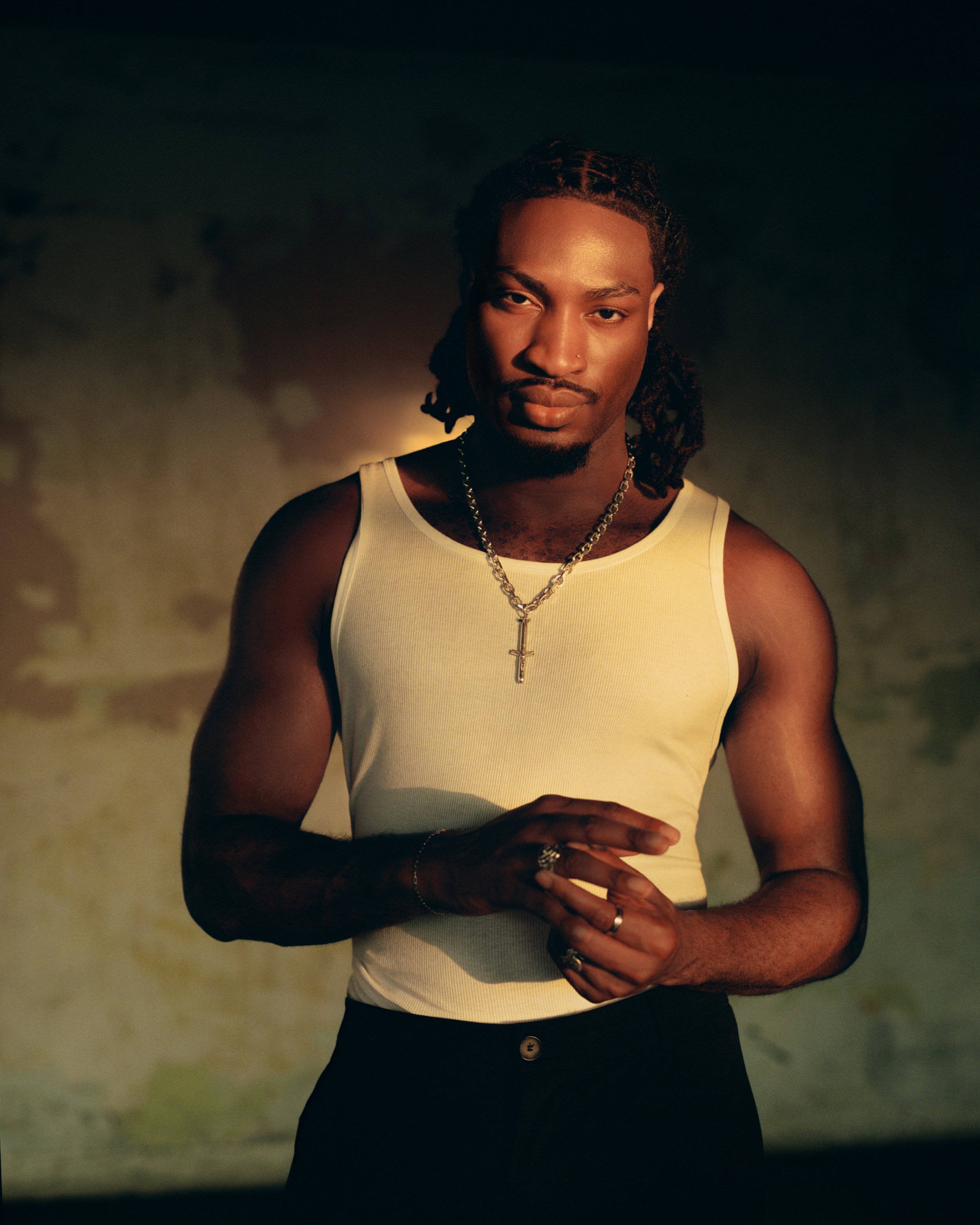 A young man with dreadlocks, wearing a white tank top and jewelry, standing on a beach at sunset.