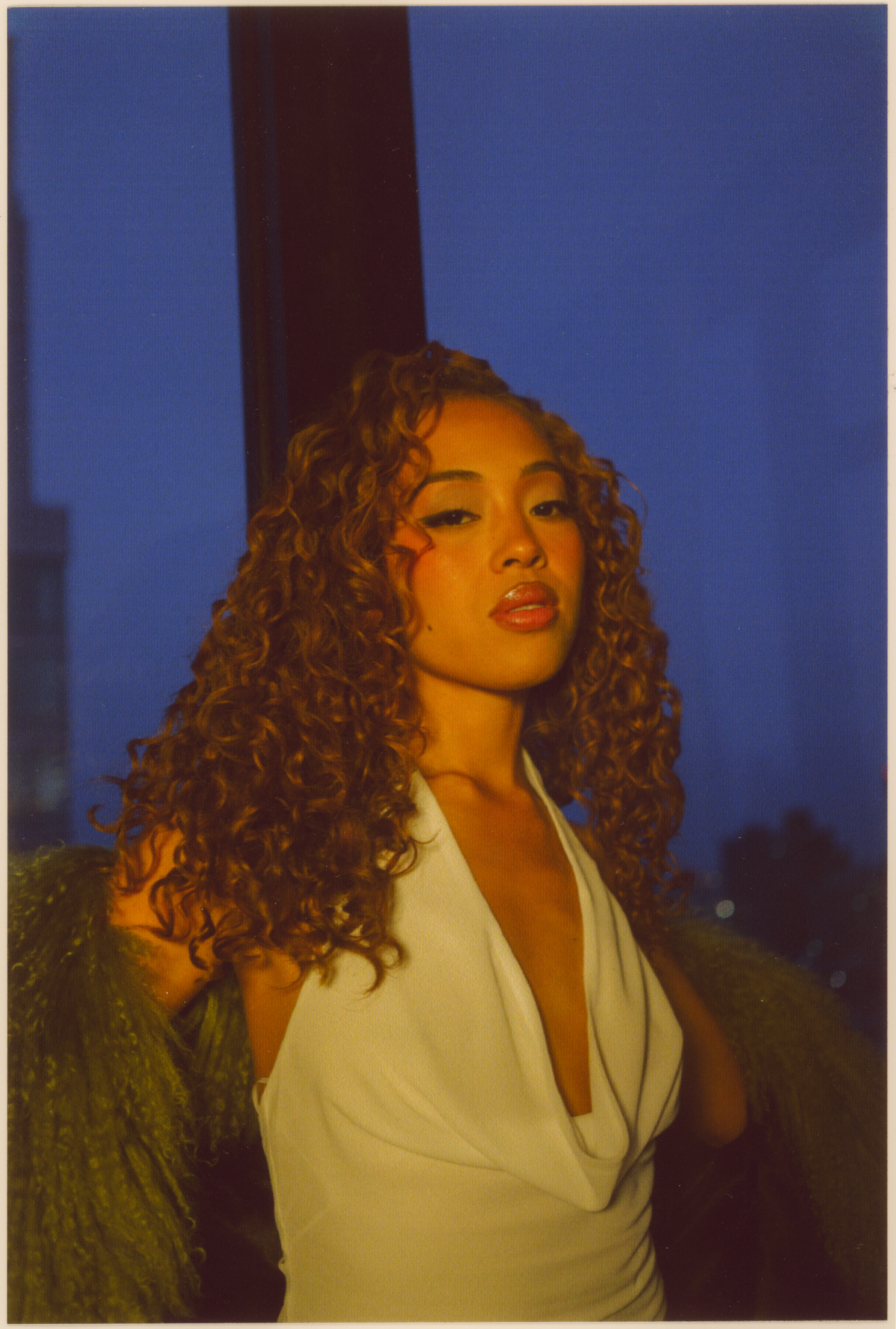 A woman with curly hair wearing a white top and a green shawl, standing outdoors at dusk with city lights in the background.