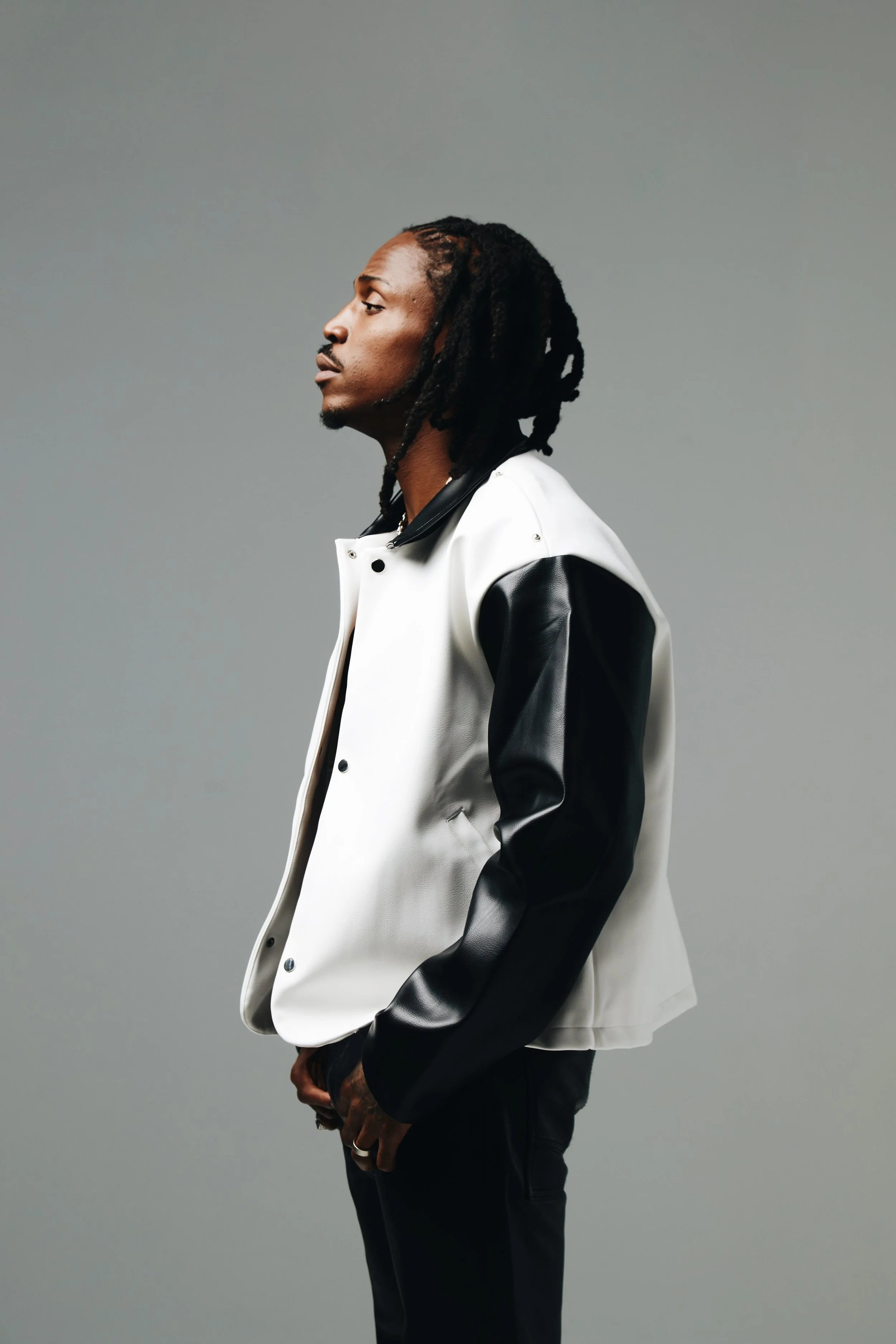 A man with dreadlocks wearing a white and black varsity jacket standing against a gray background.