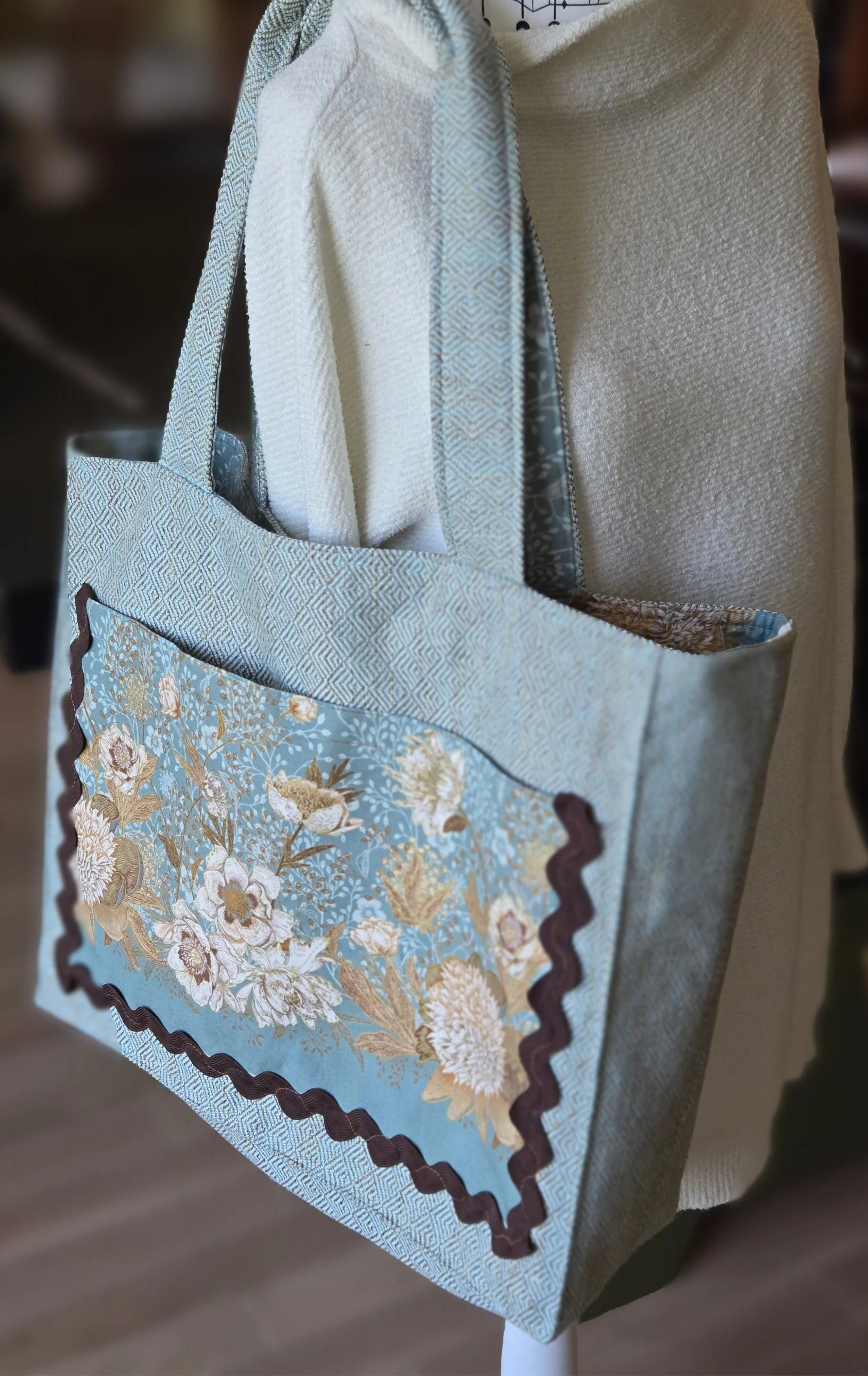 A decorative fabric tote bag is light blue with a floral pattern and has a dark brown wavy trim along the edges. exclusive to dbfourdesign