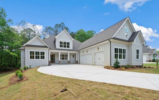 Modern white house with a gray roof, large windows, and a three-car garage, surrounded by a landscaped yard and trees under a partly cloudy sky.