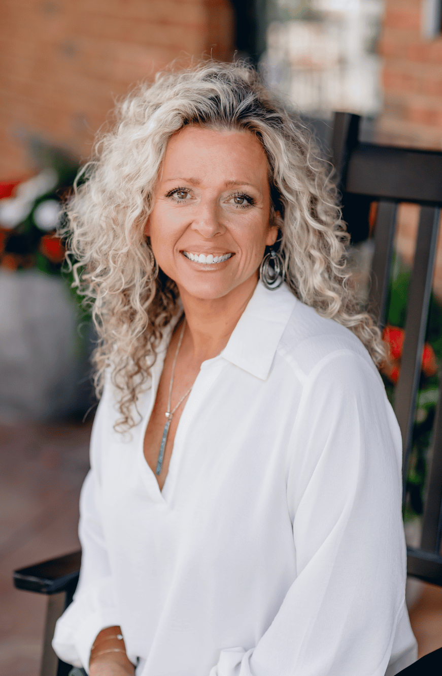 A woman with curly blonde hair smiling outdoors, wearing a white blouse and jewelry.