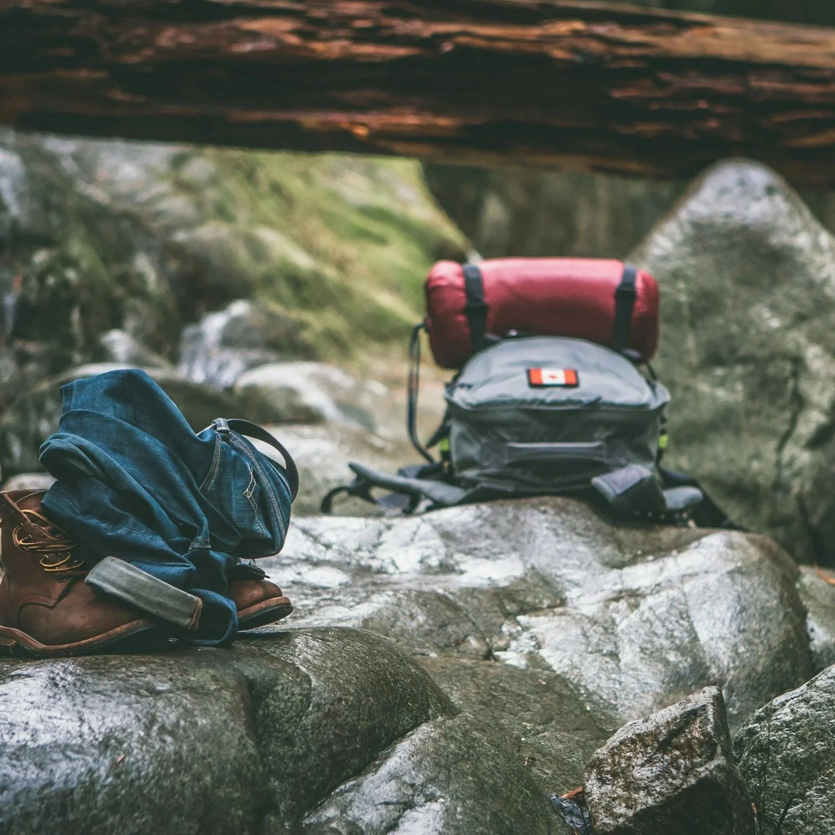 Close-up of a person’s brown hiking boots with blue jeans, resting on wet rocks near a stream surrounded by green moss-covered rocks and a natural outdoor setting.
